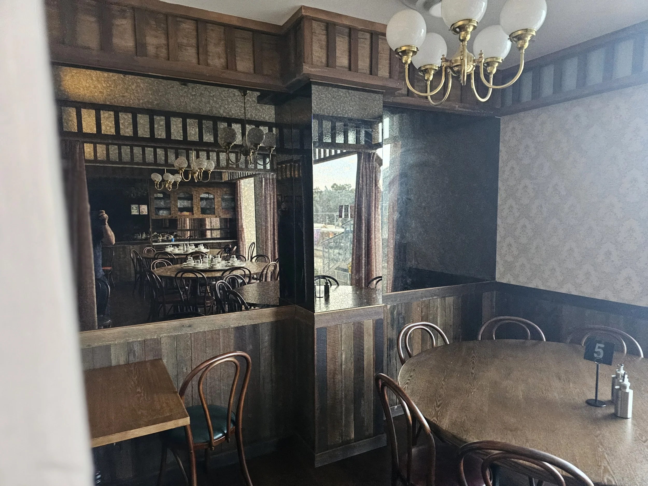 Empty restaurant dining area with wooden tables and chairs, decorative wall panels, floral curtains, vintage chandelier, and sunlight coming through large windows.