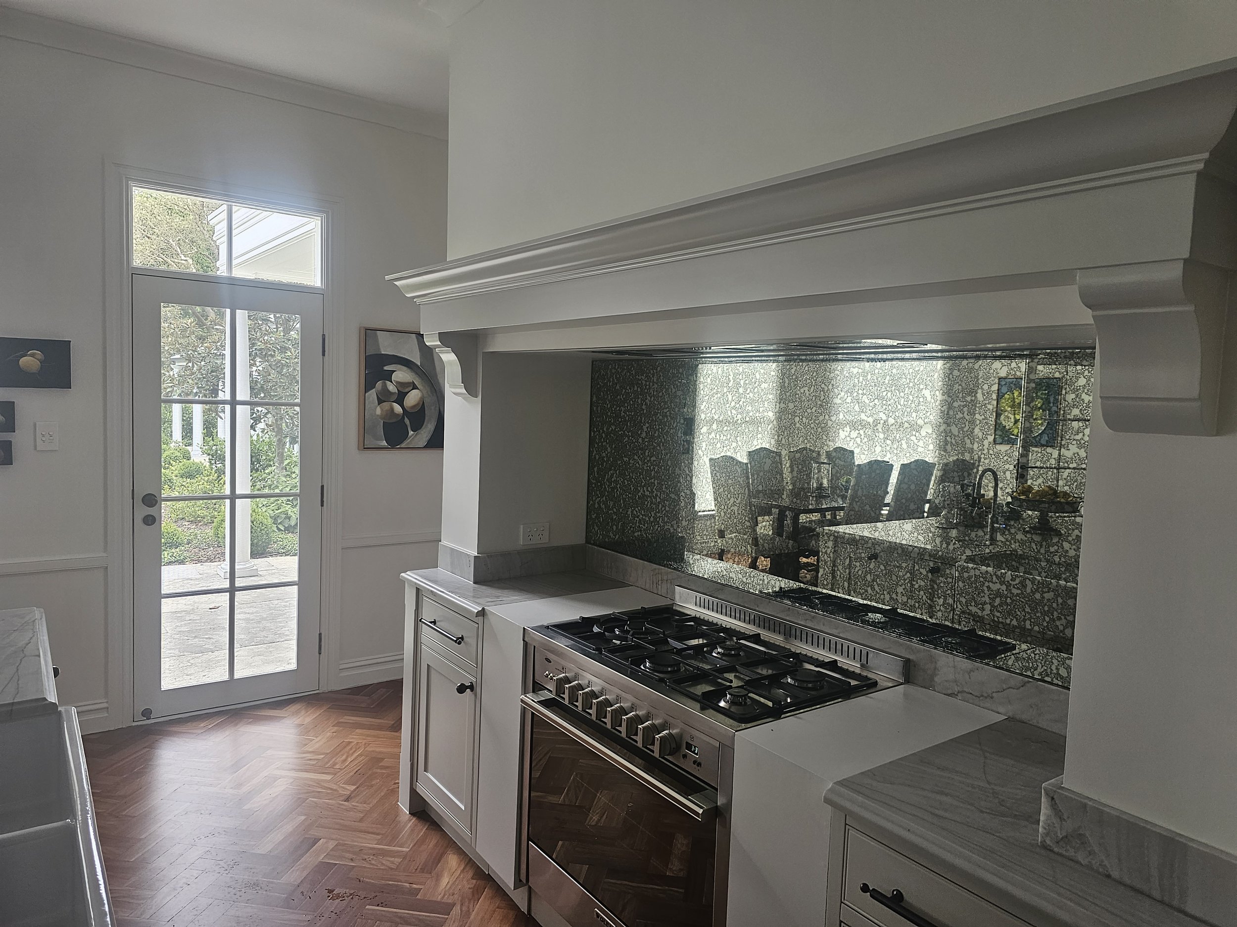 A modern kitchen with white cabinets, a stainless steel stove, marble countertops, hardwood flooring, and a glass door leading outside. There is a mirror behind the stove reflecting the dining area and window.