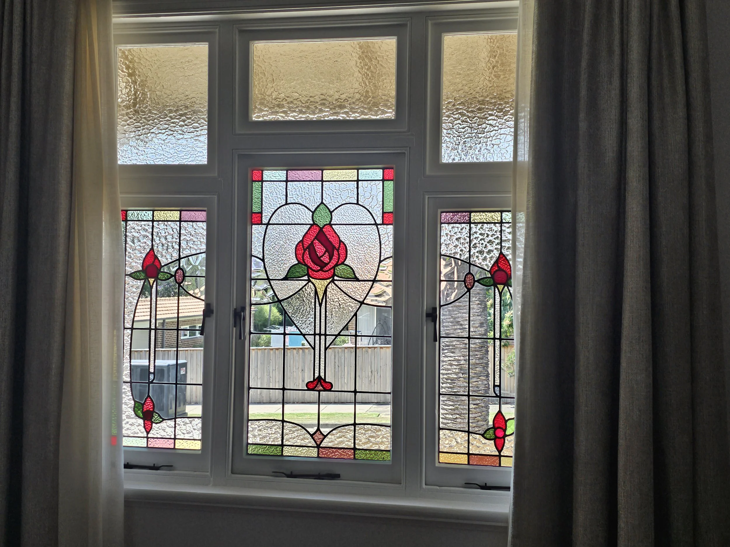 A window with stained glass featuring a central rose motif, surrounded by geometric patterns in red, green, yellow, and purple, as curtains partially frame the window.