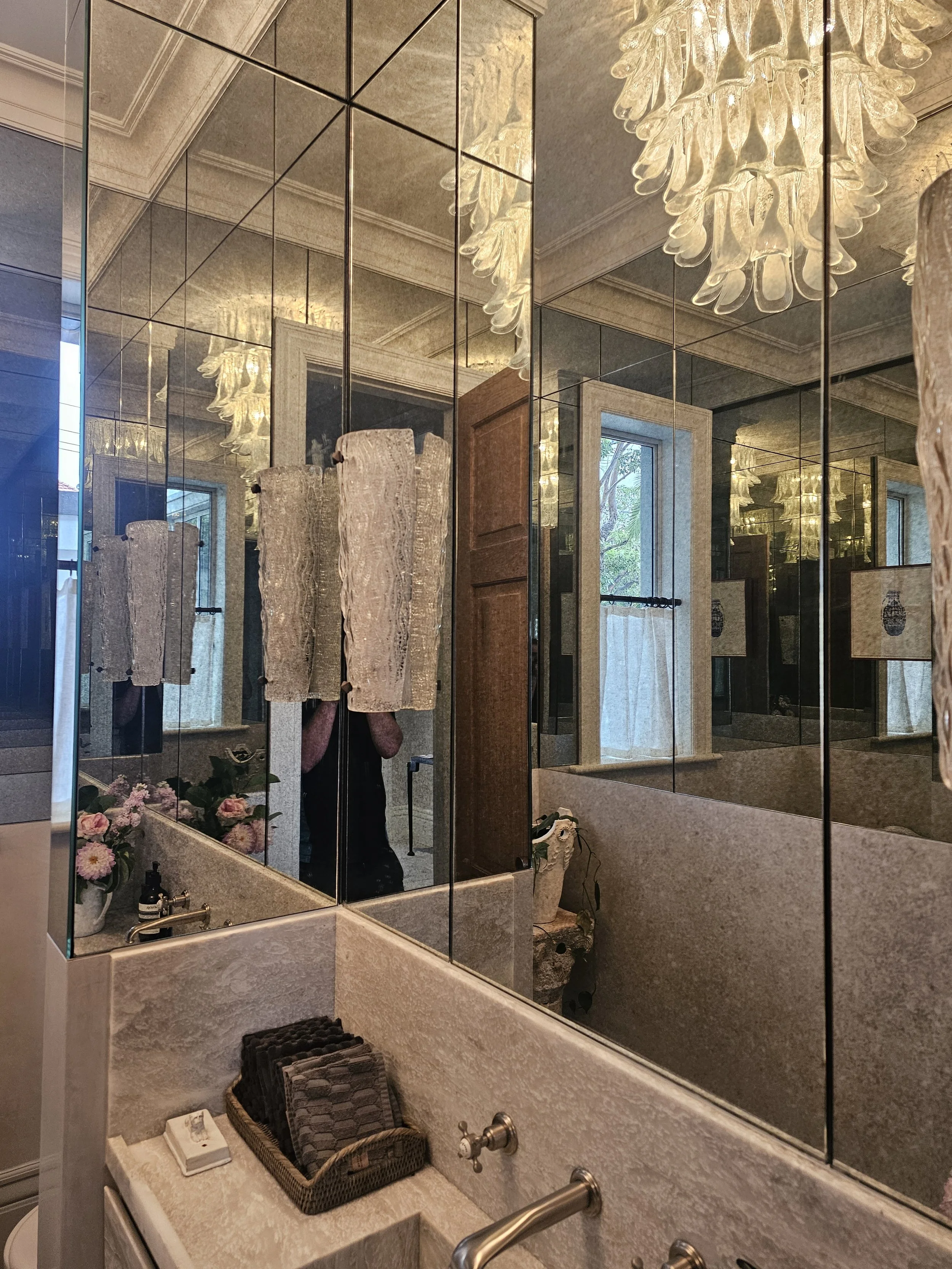 Bathroom with mirrored walls, chandelier lighting, a sink with a basket of towels, and windows with curtains, reflecting the room's decor.