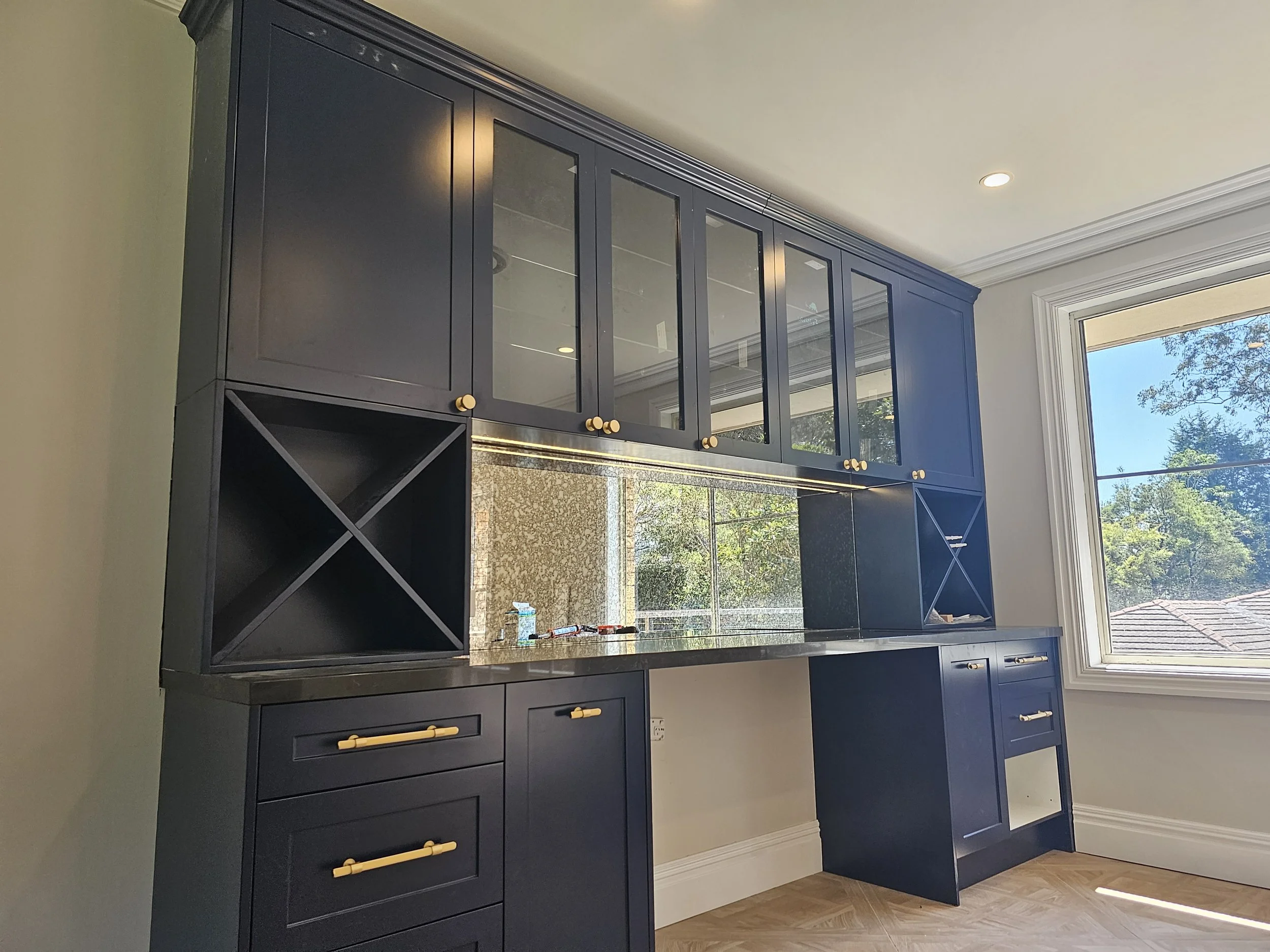 Kitchen cabinetry with navy blue cabinets, glass-paneled upper cabinets, gold handles, and wine storage sections near a window with a view of trees and a house roof outside.
