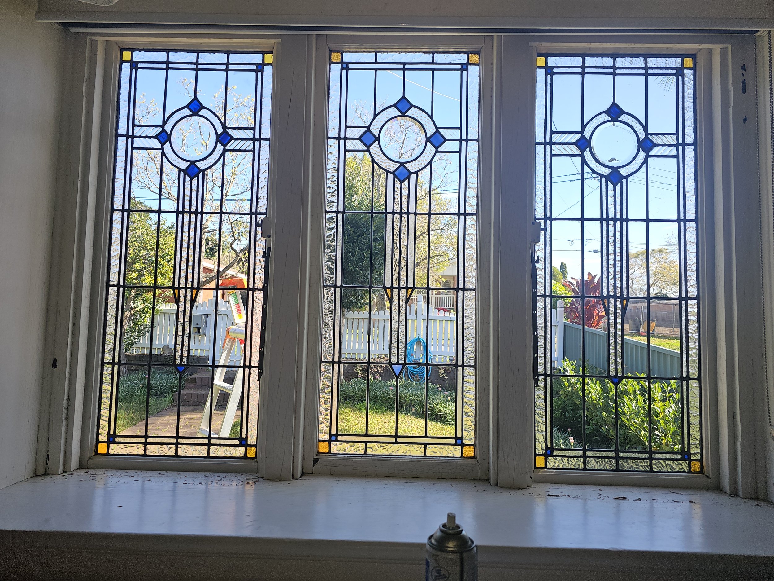 Three stained glass windows with geometric patterns looking out to a garden with trees, a white fence, and a children's slide.