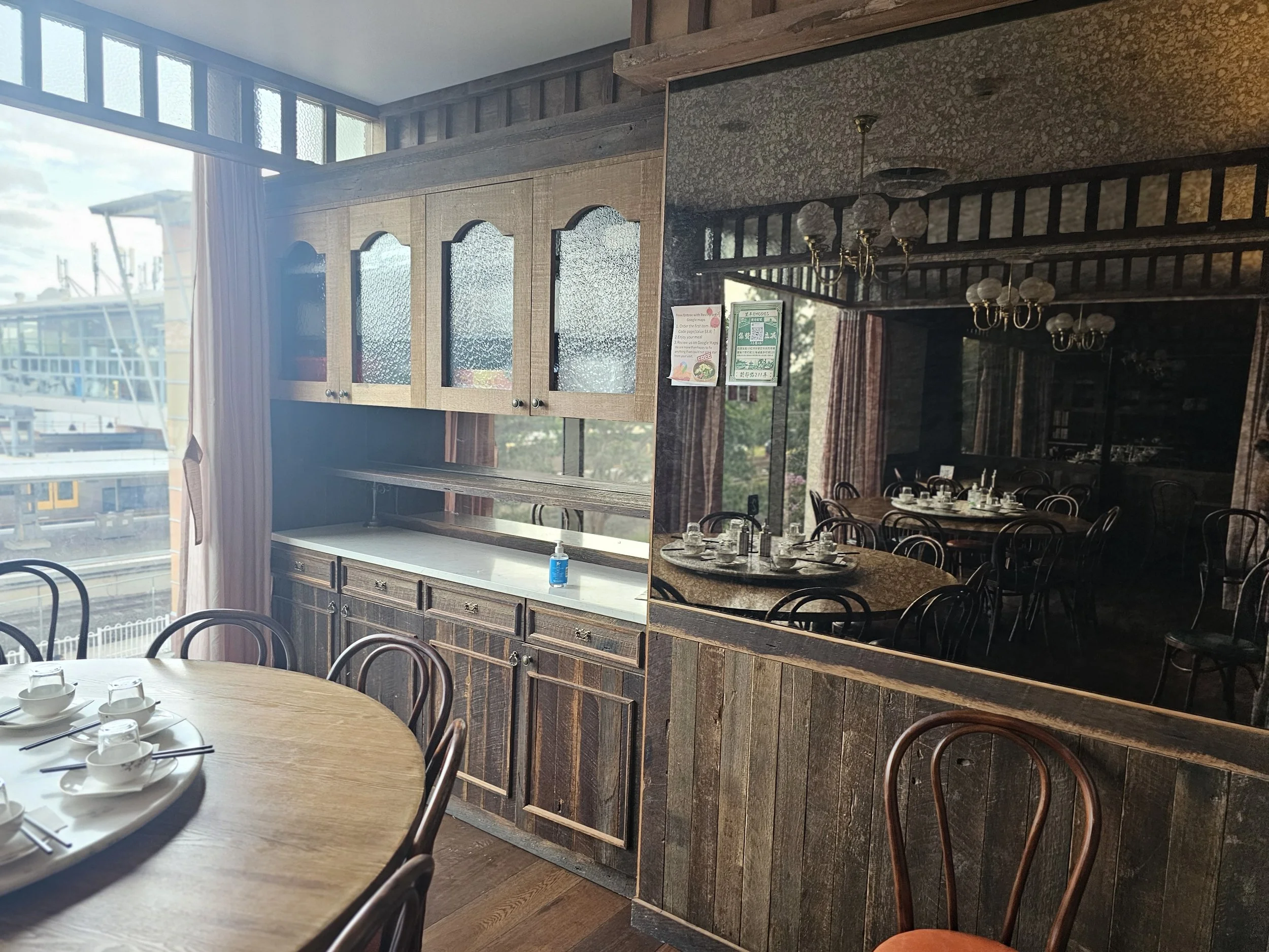 A cozy restaurant interior with a round wooden table set with white cups, saucers, and utensils, near a large window with light curtains. The room features wooden cabinetry and a wood-paneled wall, with a mirror reflecting the dining area and chandel
