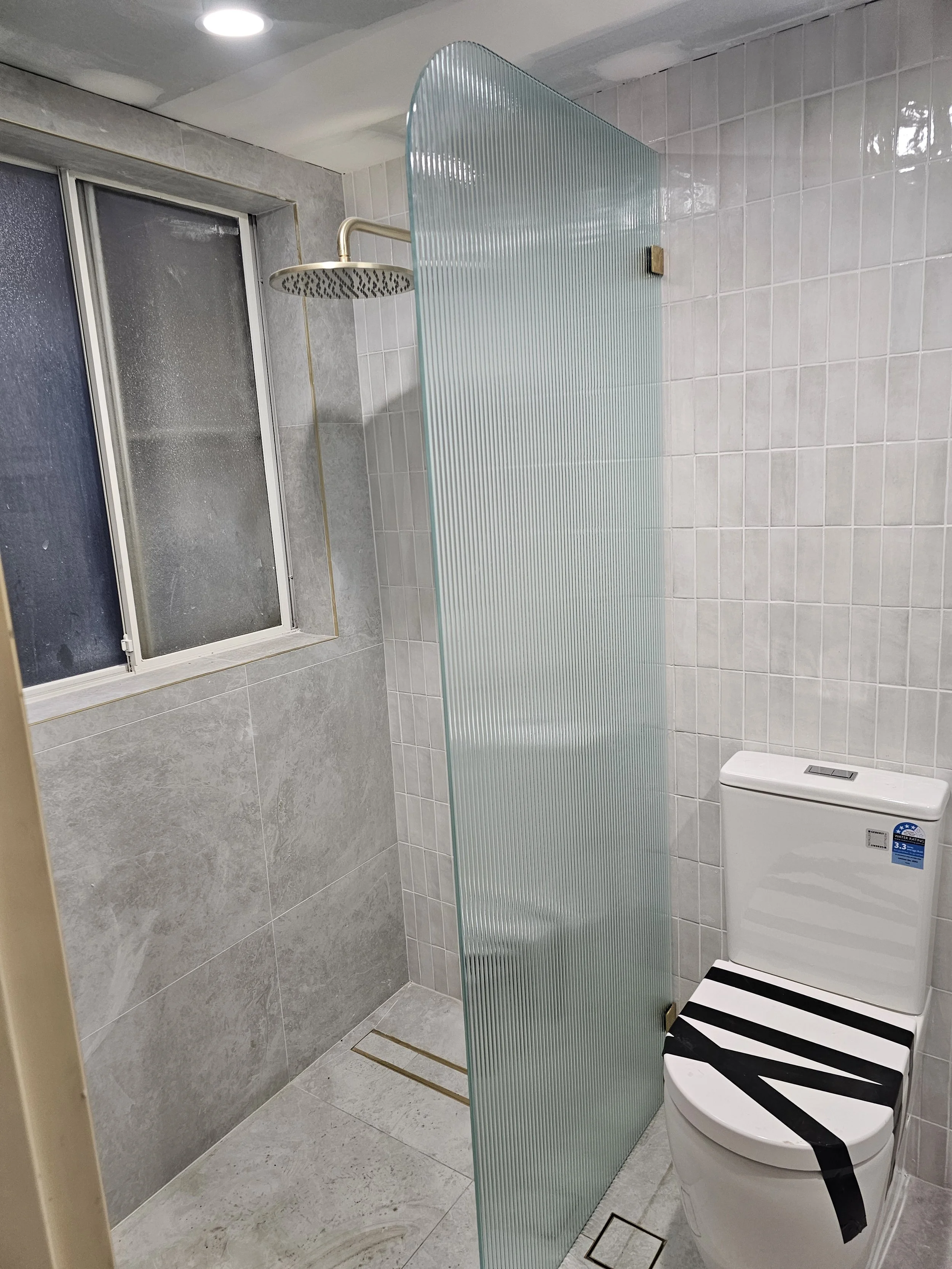 Bathroom with a bathtub, a showerhead, a frosted glass shower partition, a frosted window, and a toilet with black and white striped tape on the lid.
