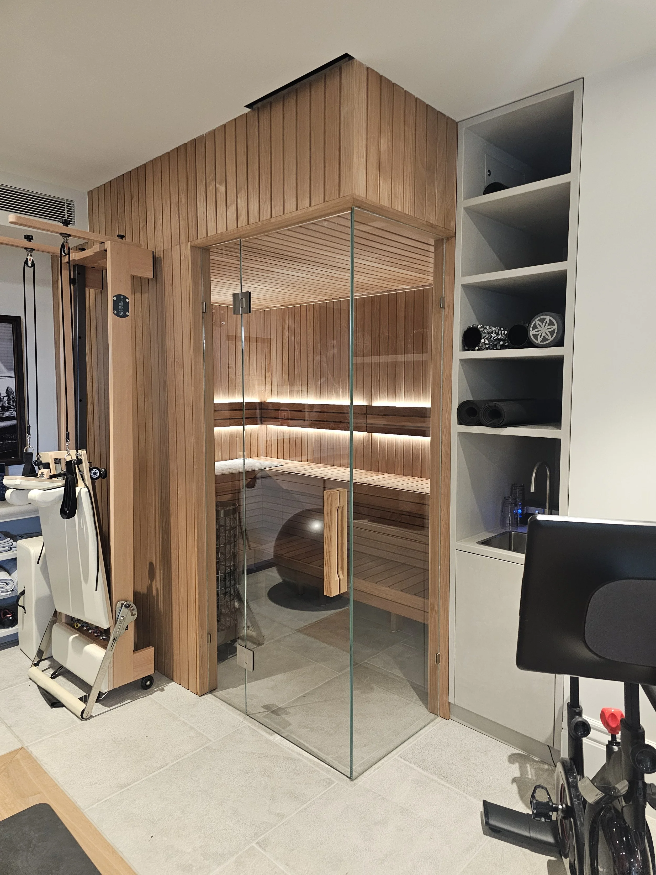 A sauna room with glass door and wooden interior, adjacent to a white storage shelf with exercise equipment, a sink, and black rolled mats.