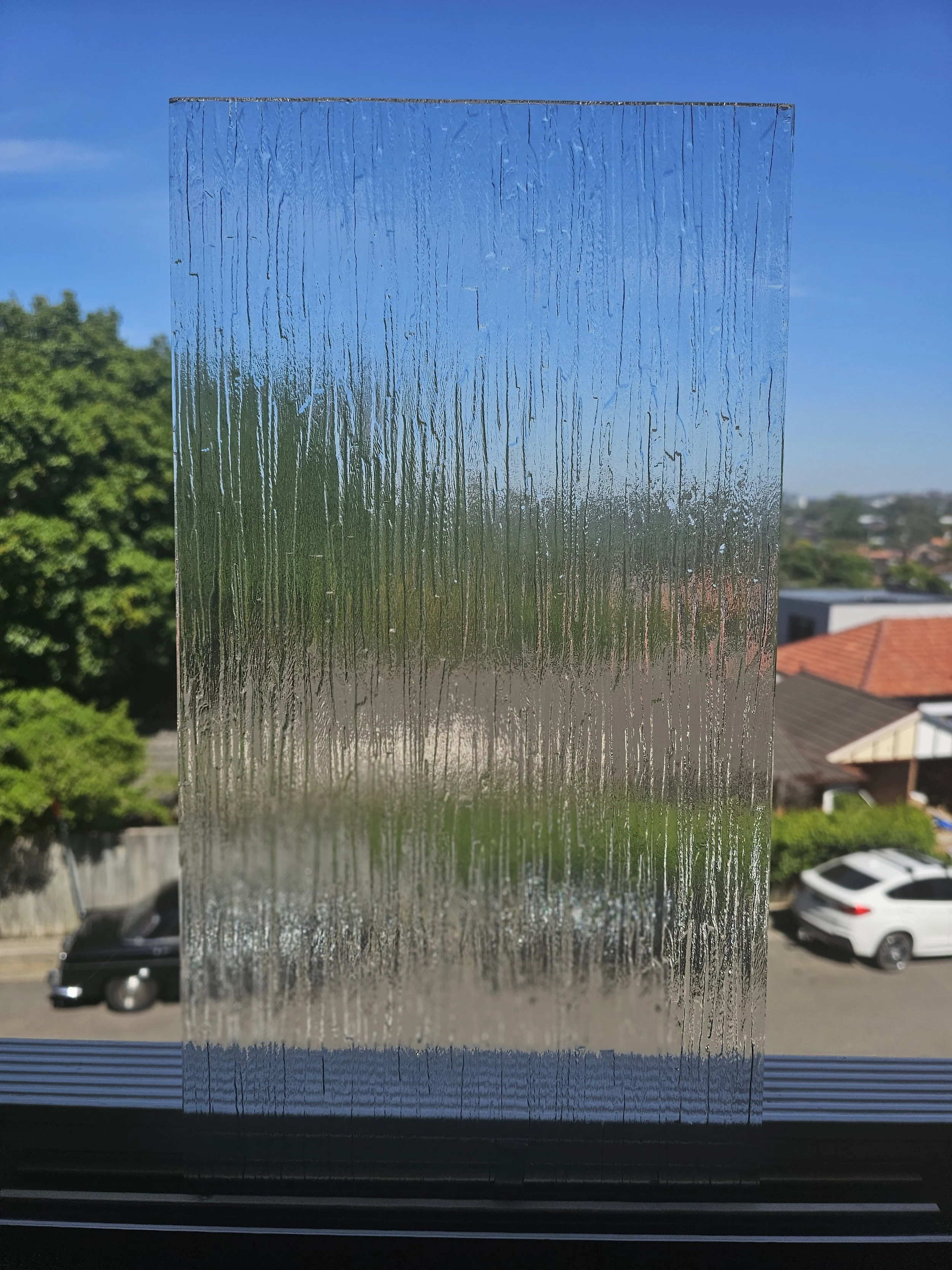 A textured window film on a window, with a view of cars, houses, trees, and a blue sky outside.