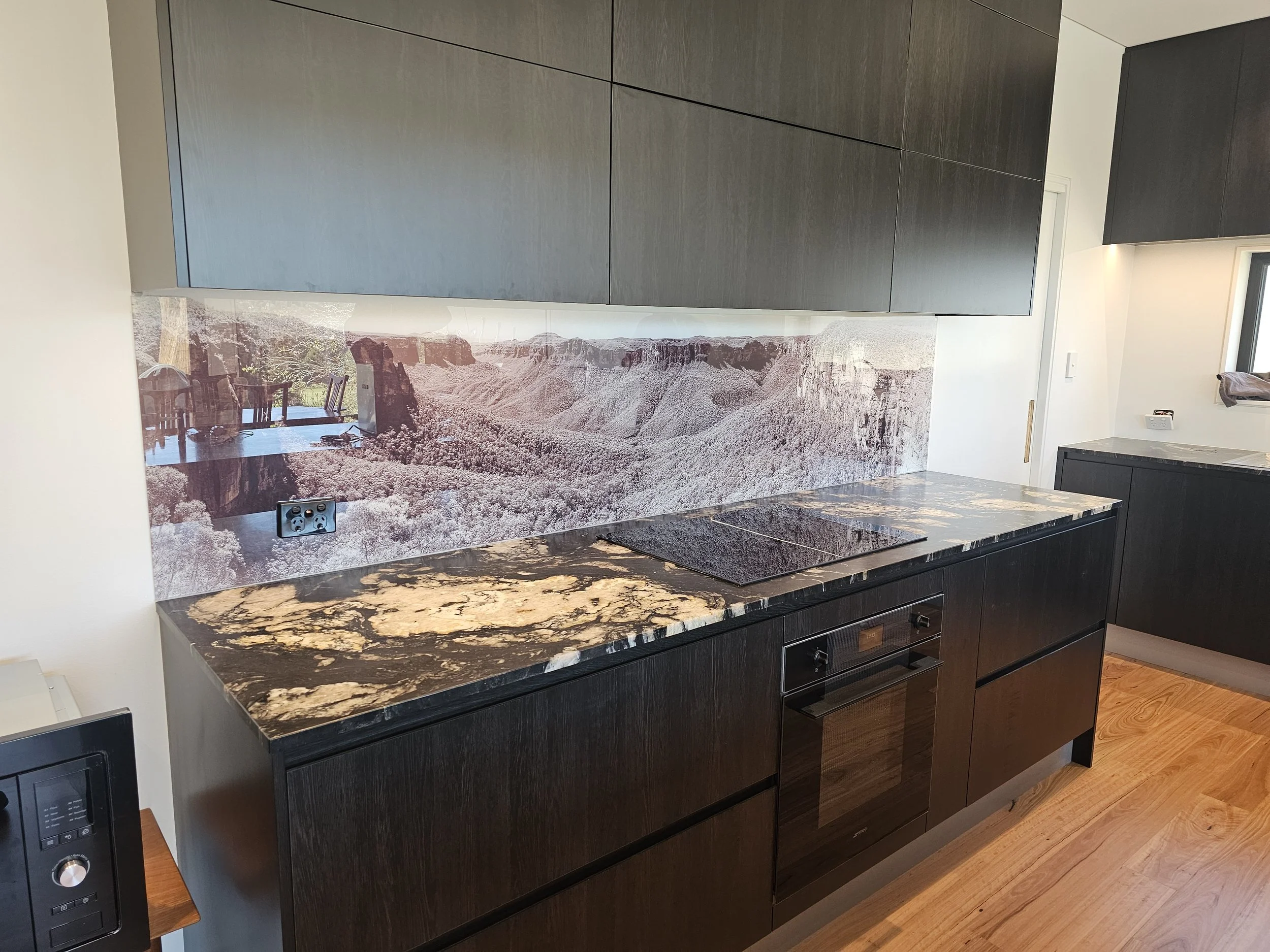 Modern kitchen with black cabinets, a black and gold marble countertop, tile backsplash with mountain landscape, and a black cooktop.