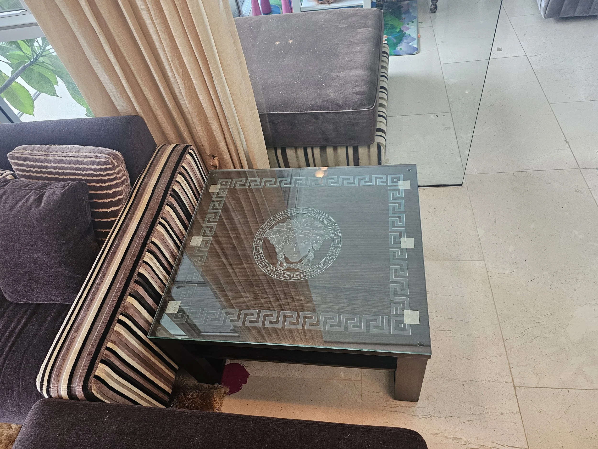 A glass coffee table with an engraved Versace Medusa logo and Greek key border, surrounded by dark and striped upholstered sofas with cushions, in a bright room with large window and beige curtains.