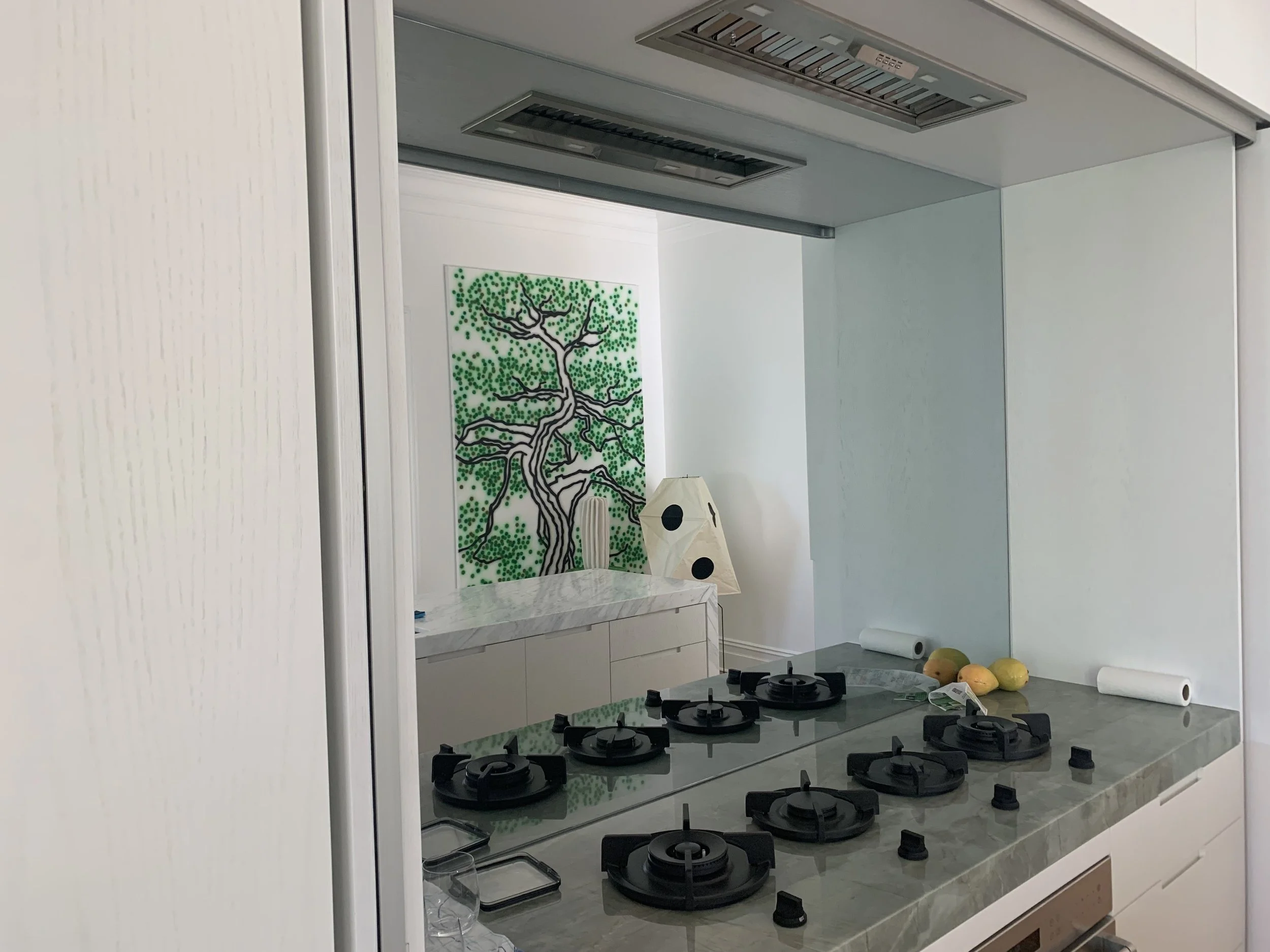 Modern kitchen with a marble countertop, black gas burners, a white artistic lamp with black circles, and a colorful tree painting on the wall.