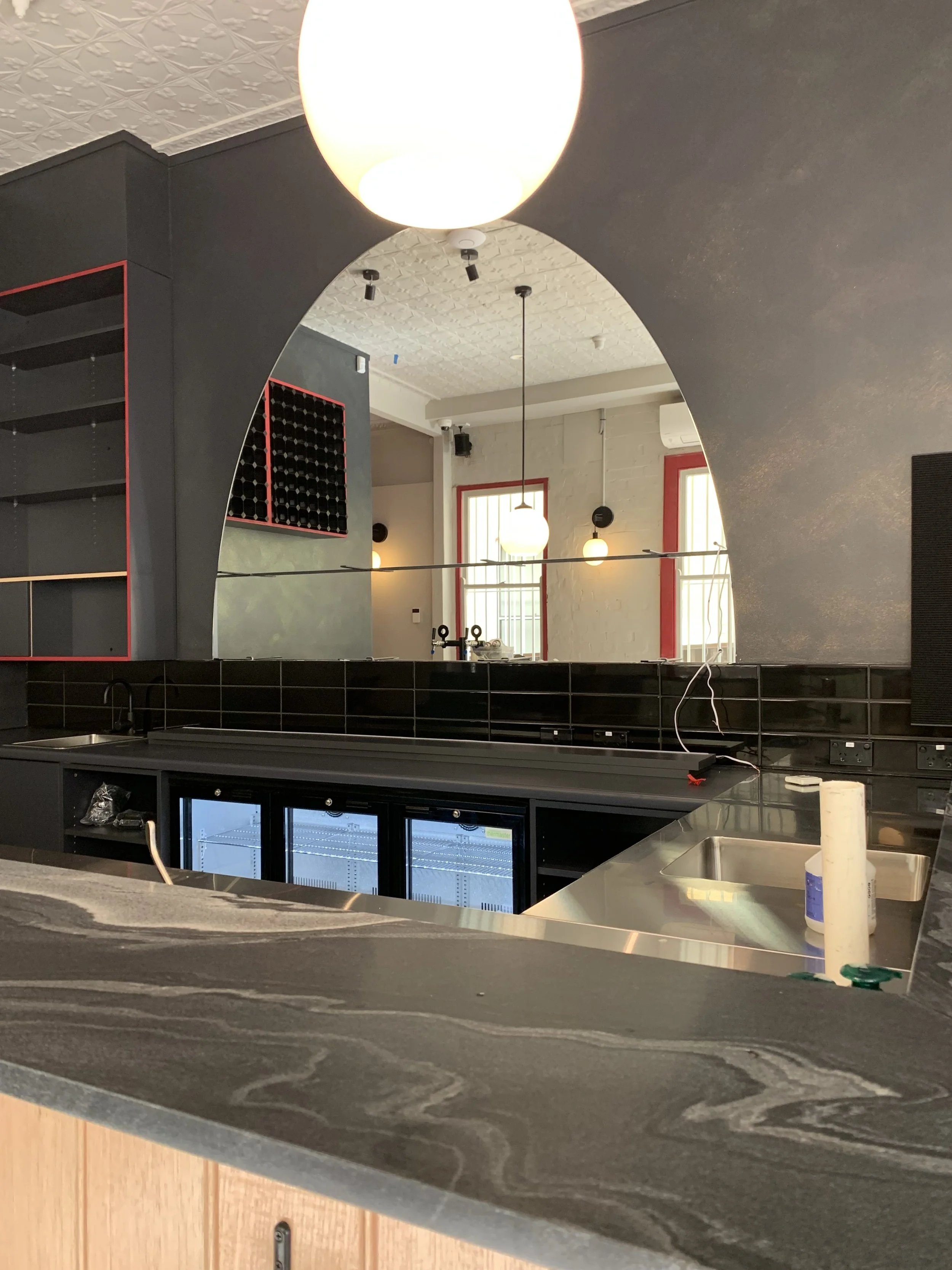 Interior of a modern bar or restaurant with black and red accents, including shelves, wine rack, and a countertop with a small sink. There are hanging pendant lights and a large mirror reflecting the seating area with large windows.