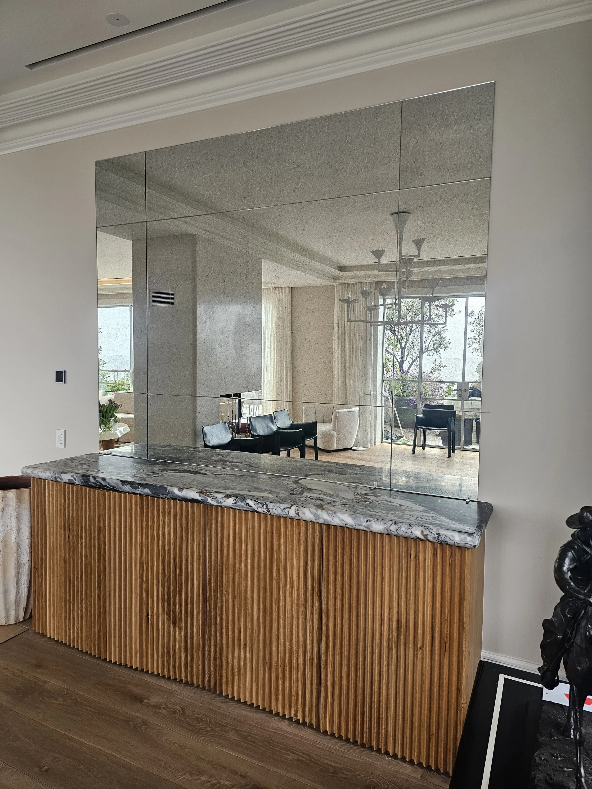 A marble-topped wooden sideboard with vertical slats, a large mirror on the wall behind it, and part of a black sculpture and framed picture on a table in foreground.