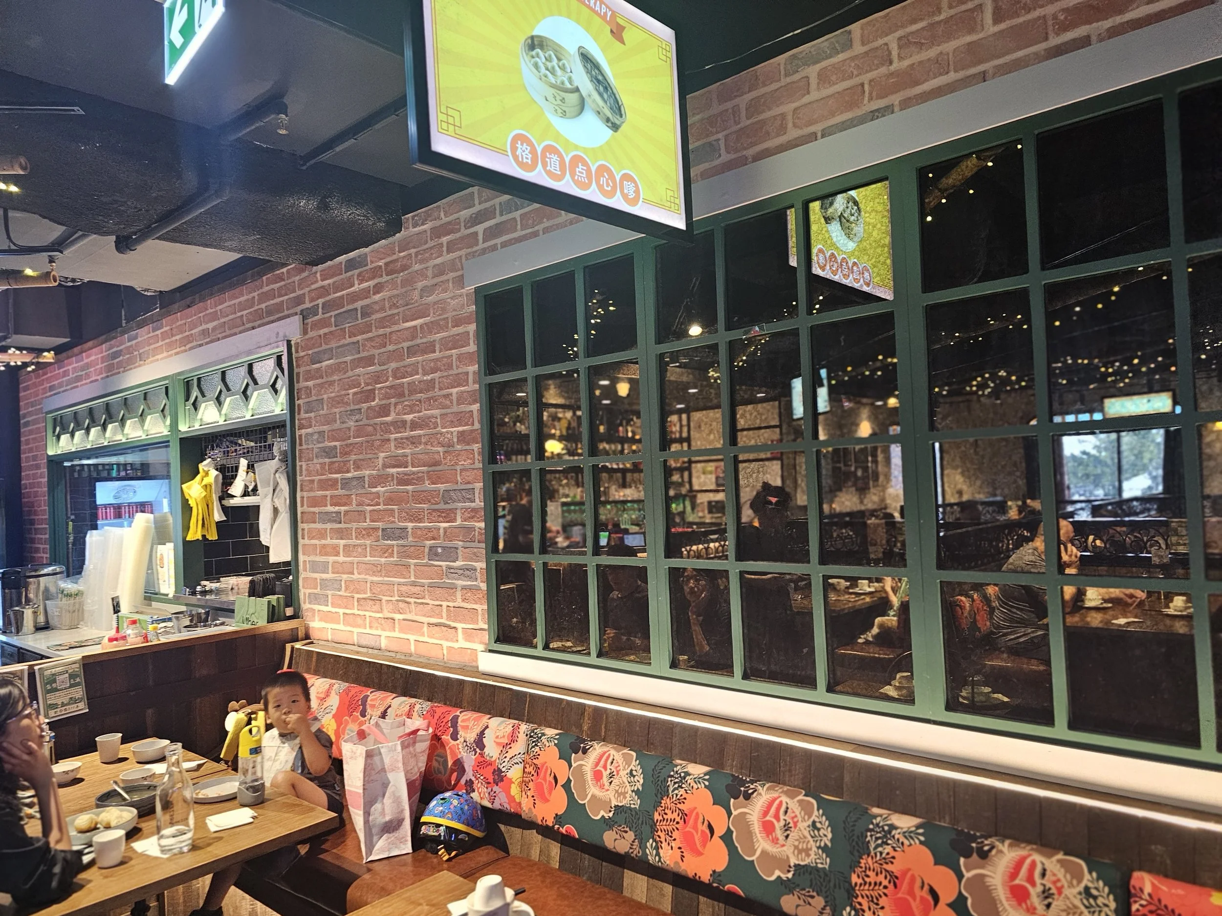 Inside a restaurant with a brick wall, a green-framed window, and a wooden dining table with dishes and a child sitting at the table. There is a television screen hanging from the ceiling showing an image of food, and some staff and customers are visible inside through the window.