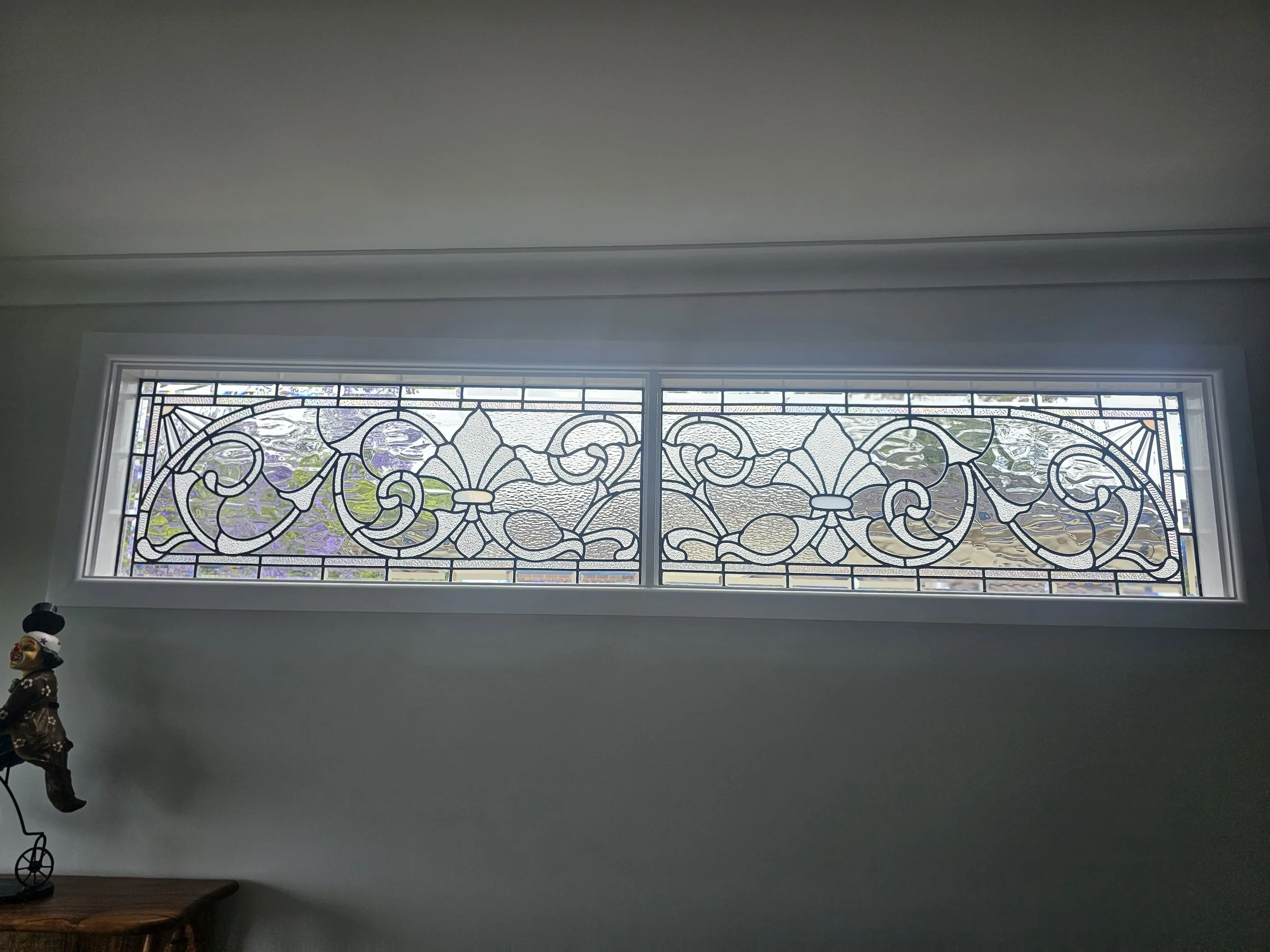 Long rectangular window with intricate floral and geometric stained glass design, overlooking an outdoor scene with trees and houses.