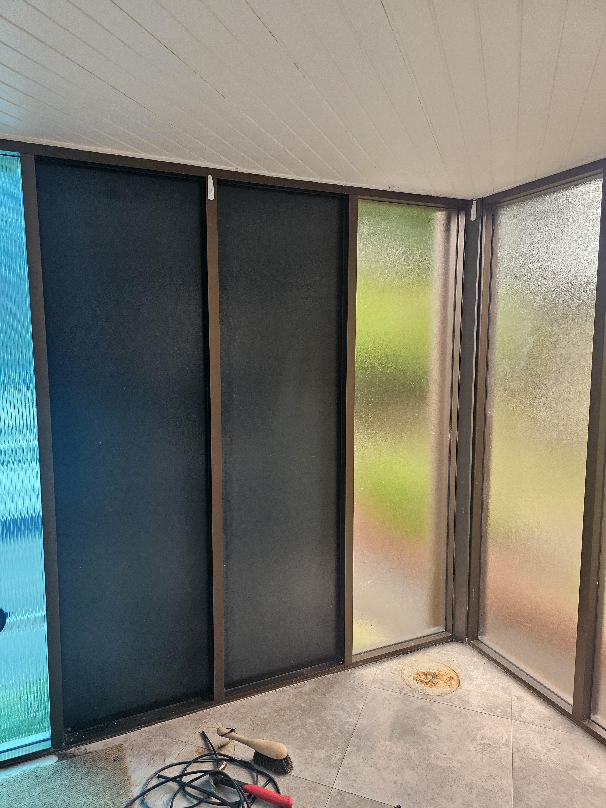 Corner of a screened-in porch with frosted glass panels, a white wood-paneled ceiling, and a tiled floor. There are cleaning tools on the ground, including a brush and a vacuum attachment.
