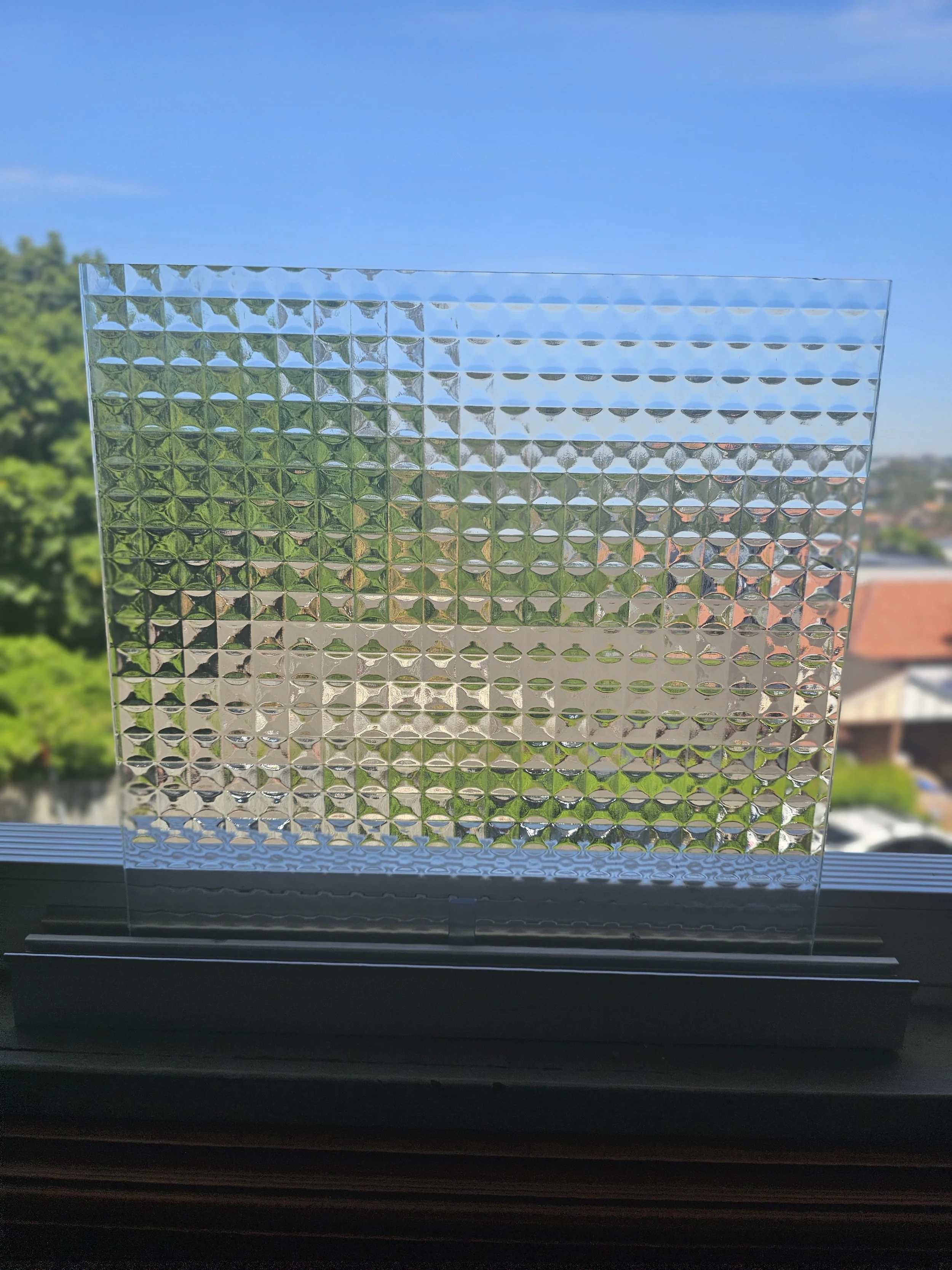 A decorative glass panel with a textured pattern, placed on a windowsill with a view of trees and houses outside on a sunny day.