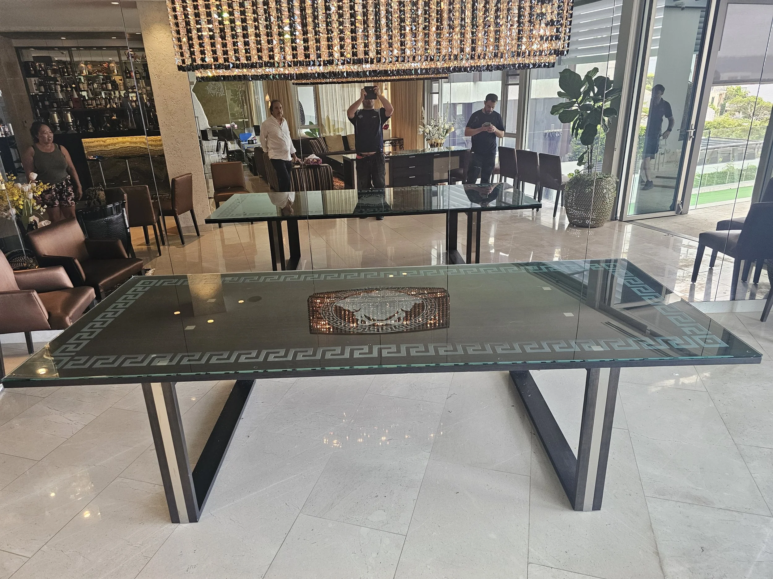 Modern dining area with glass-top table, surrounded by chairs, large windows, and people taking photos.