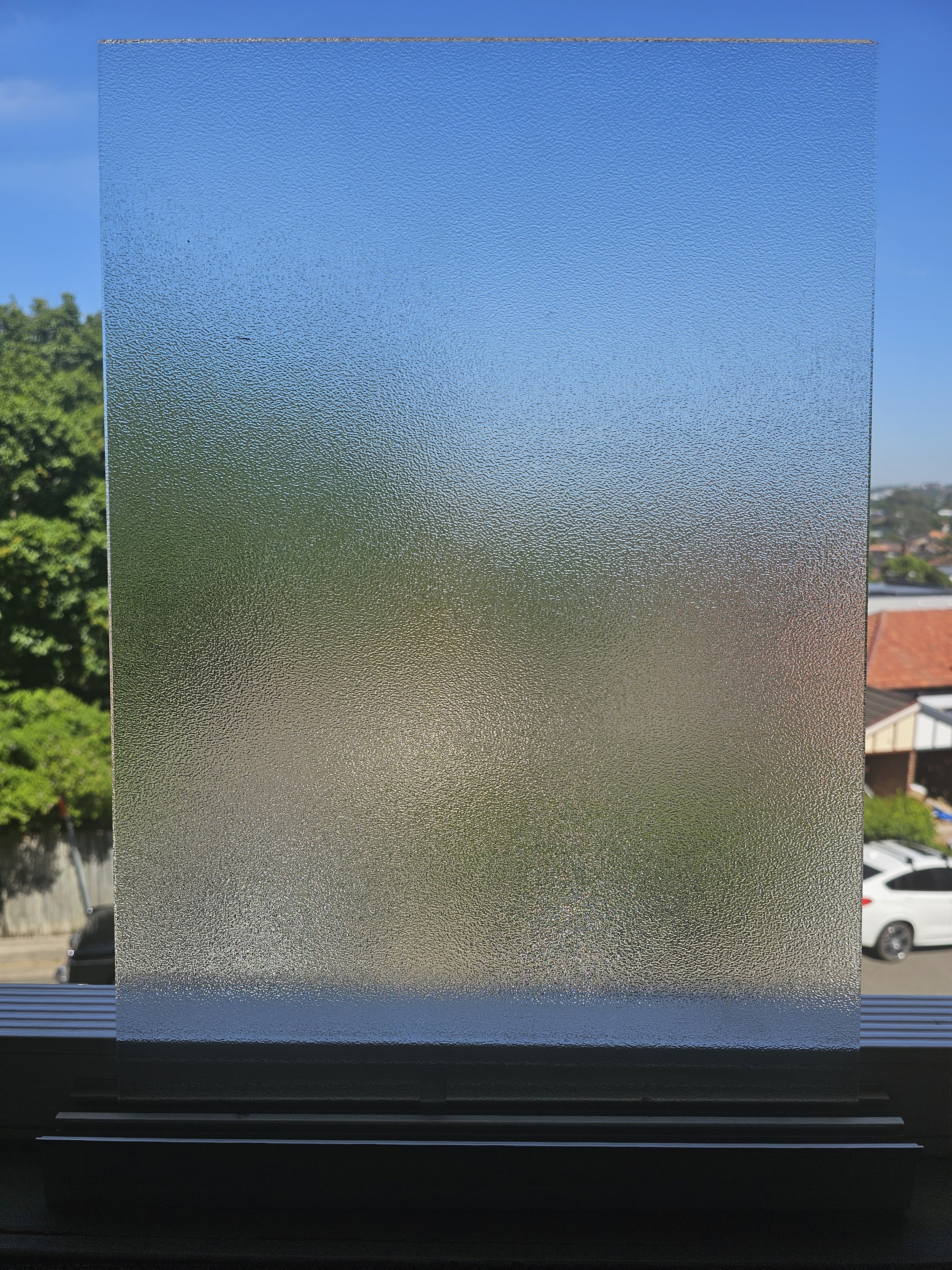 Frosted glass window with a view of trees, houses, and cars outside under a blue sky.