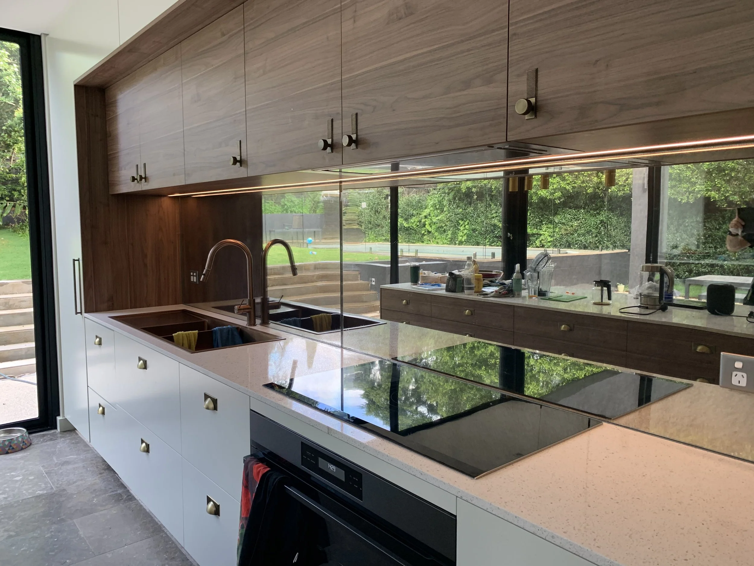 Modern kitchen with white cabinets, a speckled white countertop, a black glass cooktop, two sinks with a copper faucet, and a large mirrored backsplash reflecting a backyard with green trees and stairs.