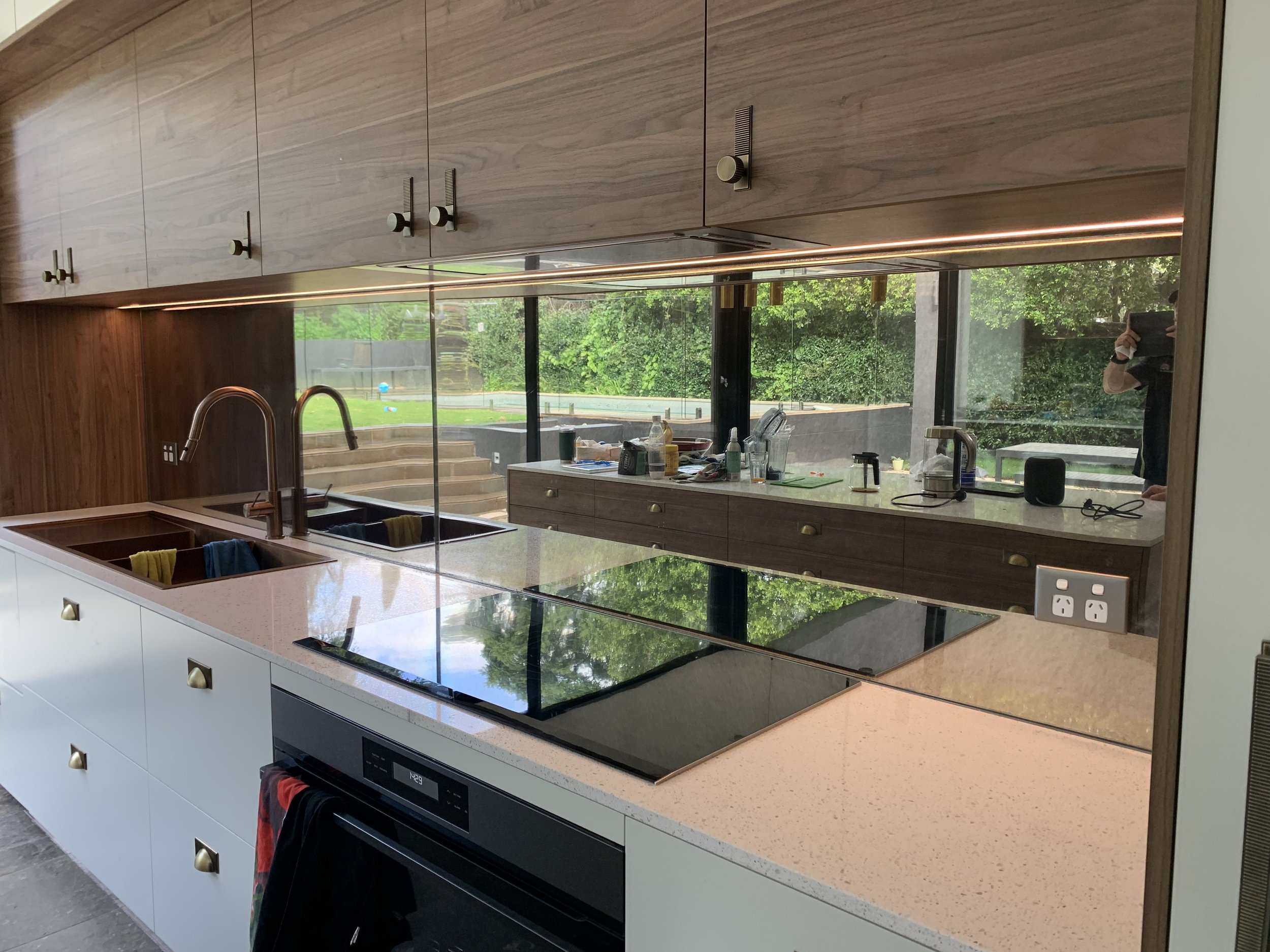 Modern kitchen with white cabinetry, a black electric cooktop, a built-in oven, and a large window reflecting the outdoors with green trees. Countertop objects include a sink, utensils, bottles, and small appliances.