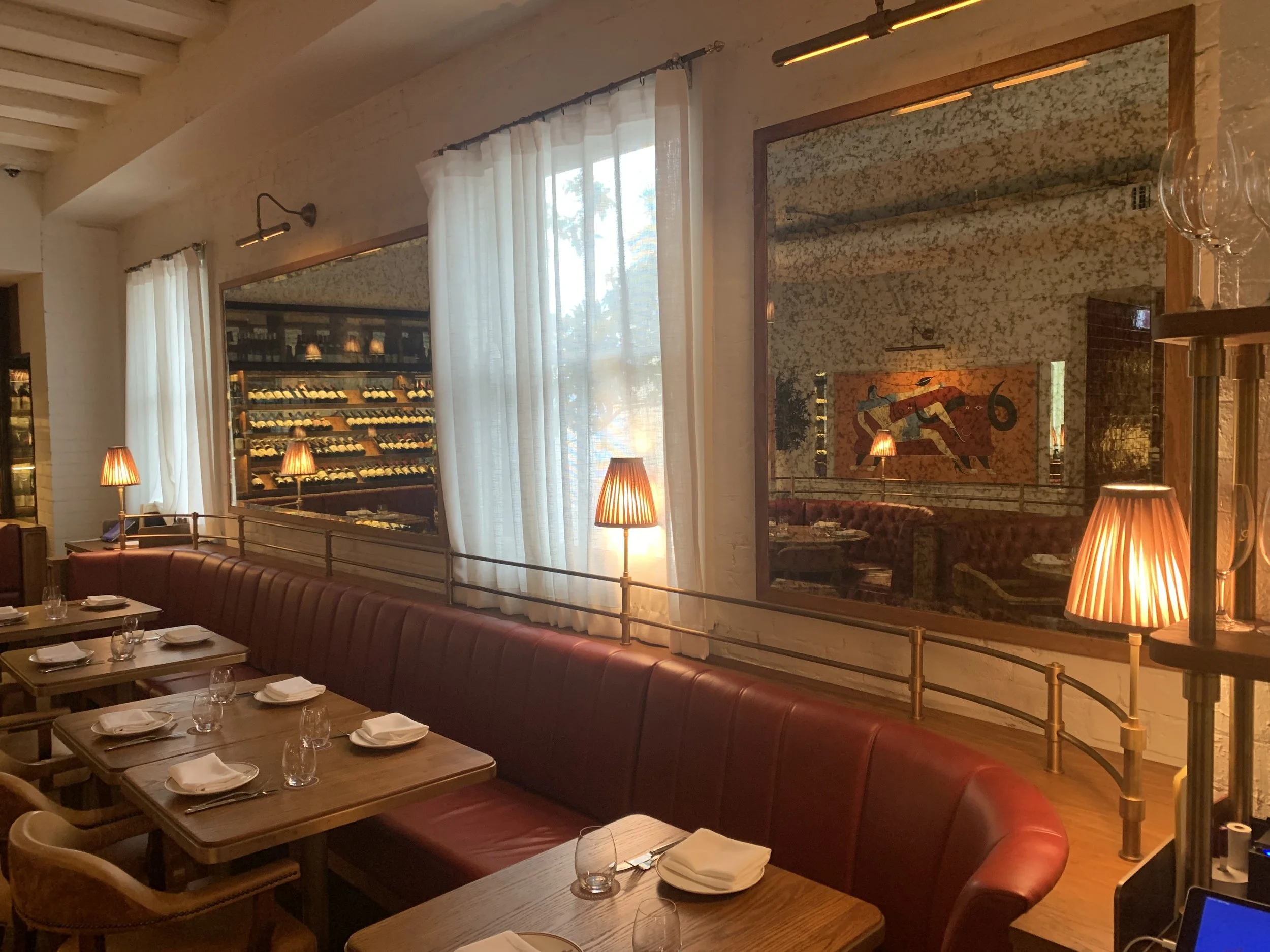 Inside a cozy restaurant with wooden tables set with napkins, glasses, and plates, featuring a red upholstered booth along the wall. Warm lighting from table lamps and wall sconces illuminates the space. Large windows with white curtains reveal a win