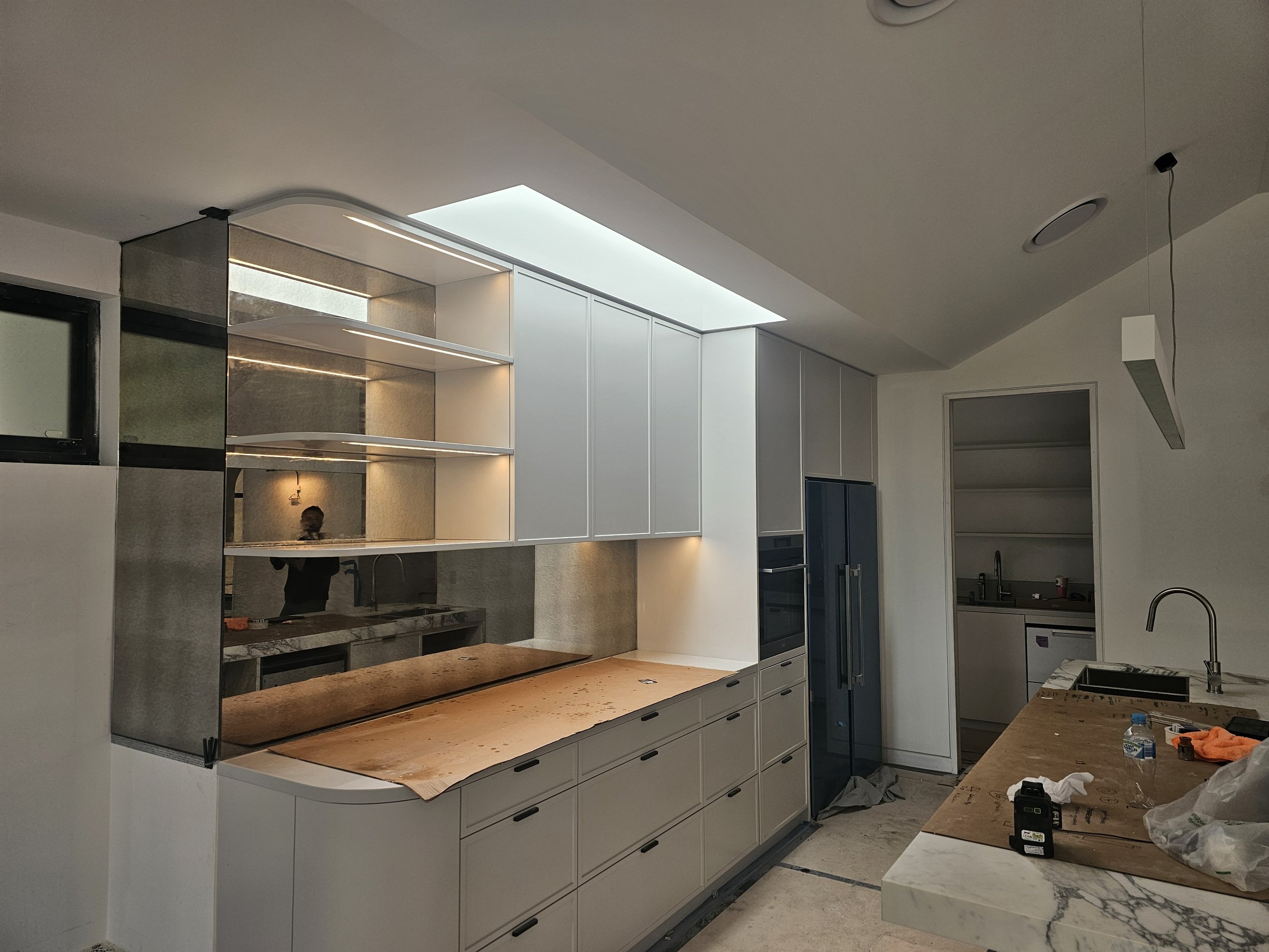 Modern kitchen under construction with white cabinets, marble countertops, a built-in oven, a refrigerator, and a skylight, with construction materials and tools on the counter.