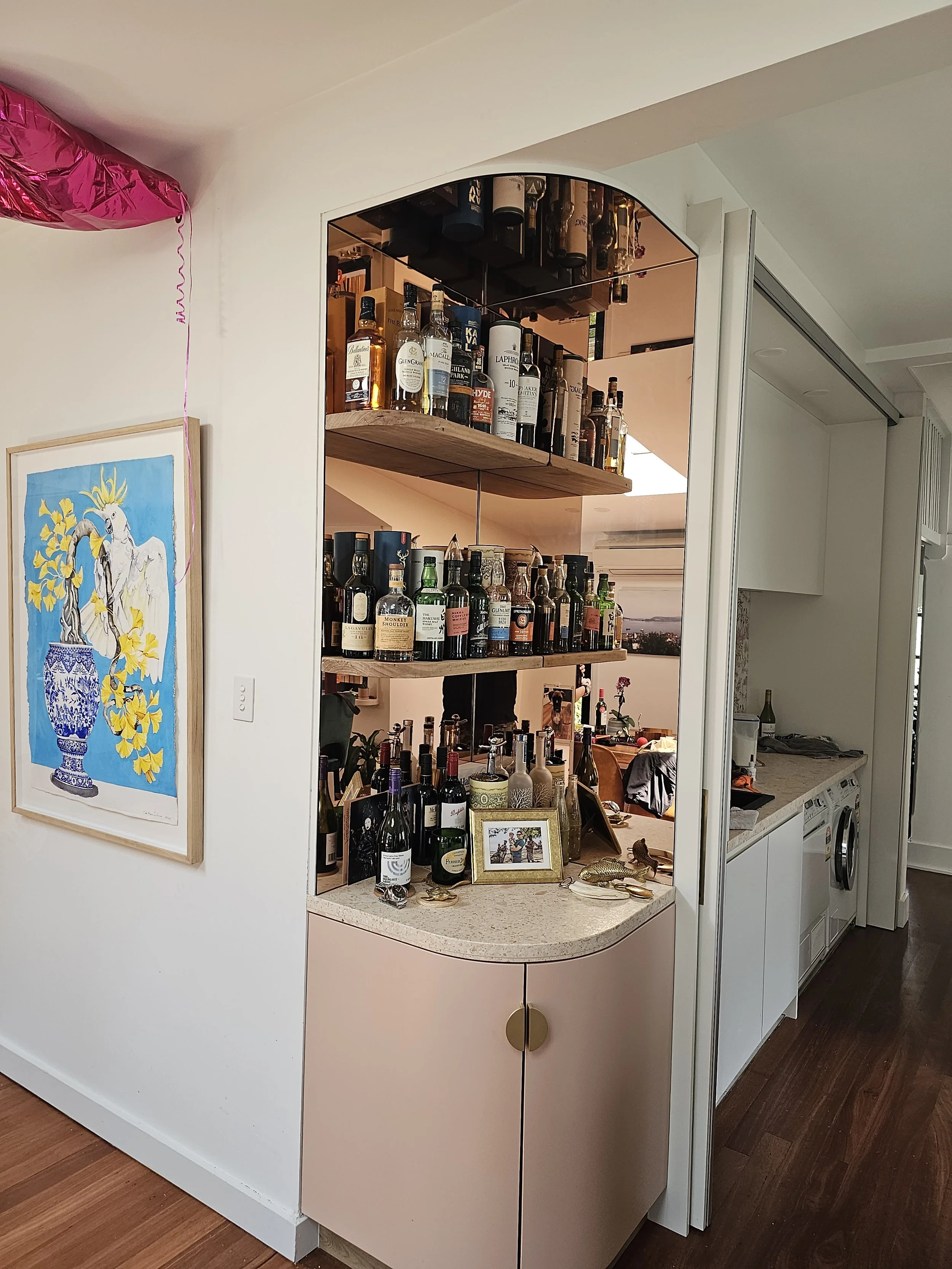 A corner of a room with a pink cabinet topped with a beige countertop, displaying various liquor bottles, a framed photo, and decorative items. Behind the bar area, there are wooden shelves filled with additional bottles. To the right, a laundry area