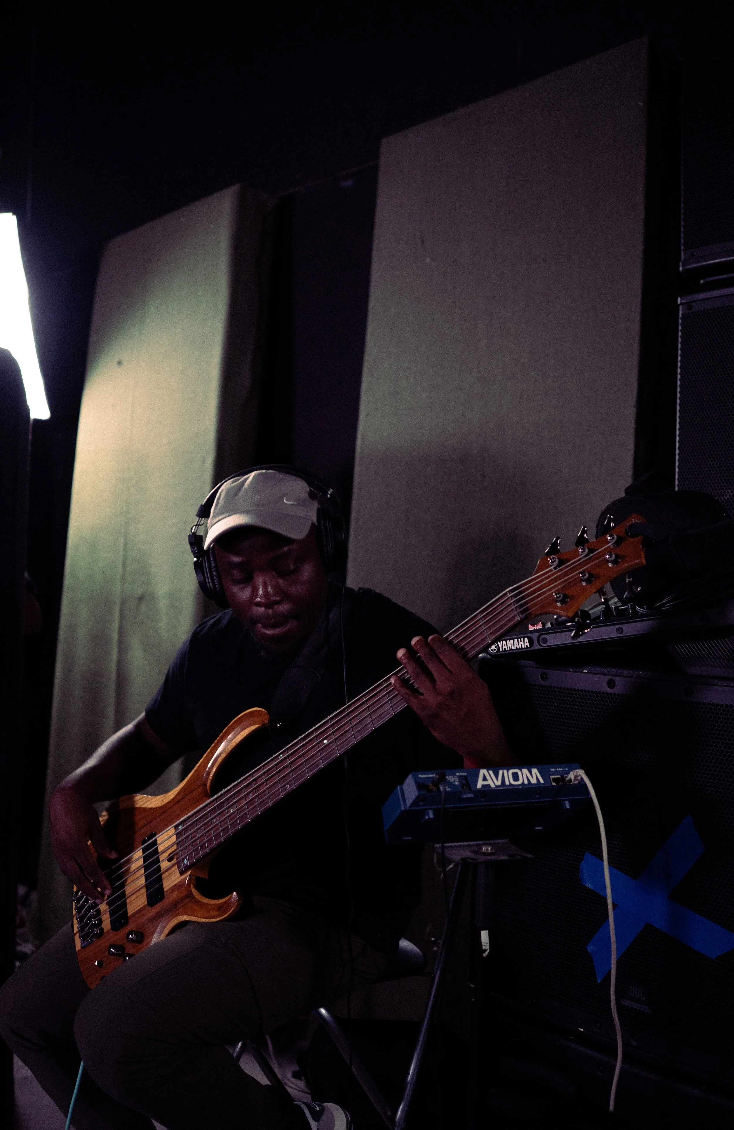 A musician wearing headphones and a beige cap playing a bass guitar in a dimly lit room with soundproofing panels and audio equipment.