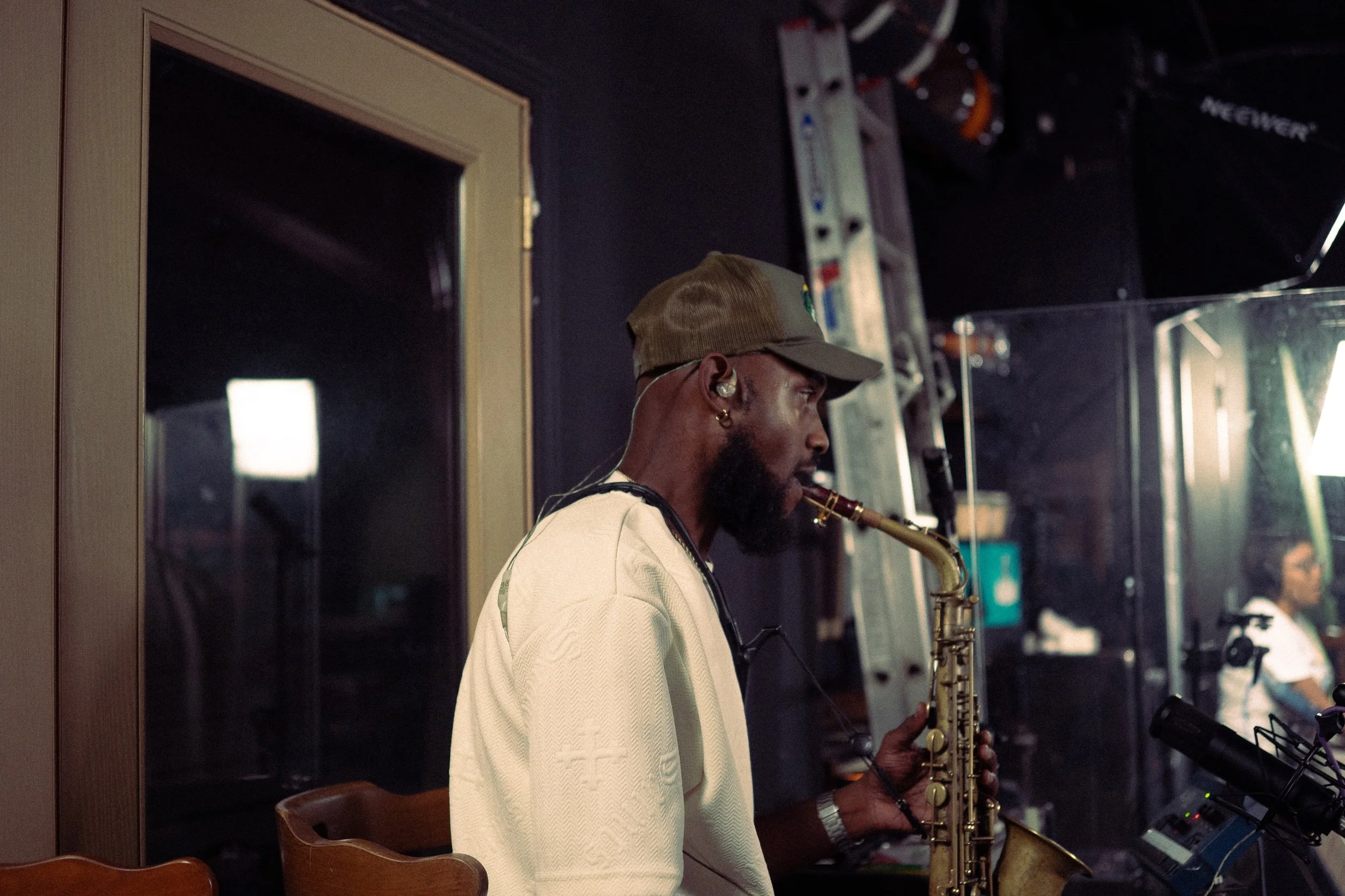 A man playing a saxophone in a recording studio, wearing a beige cap, wireless earbuds, and a white shirt, with recording equipment and a woman in the background.