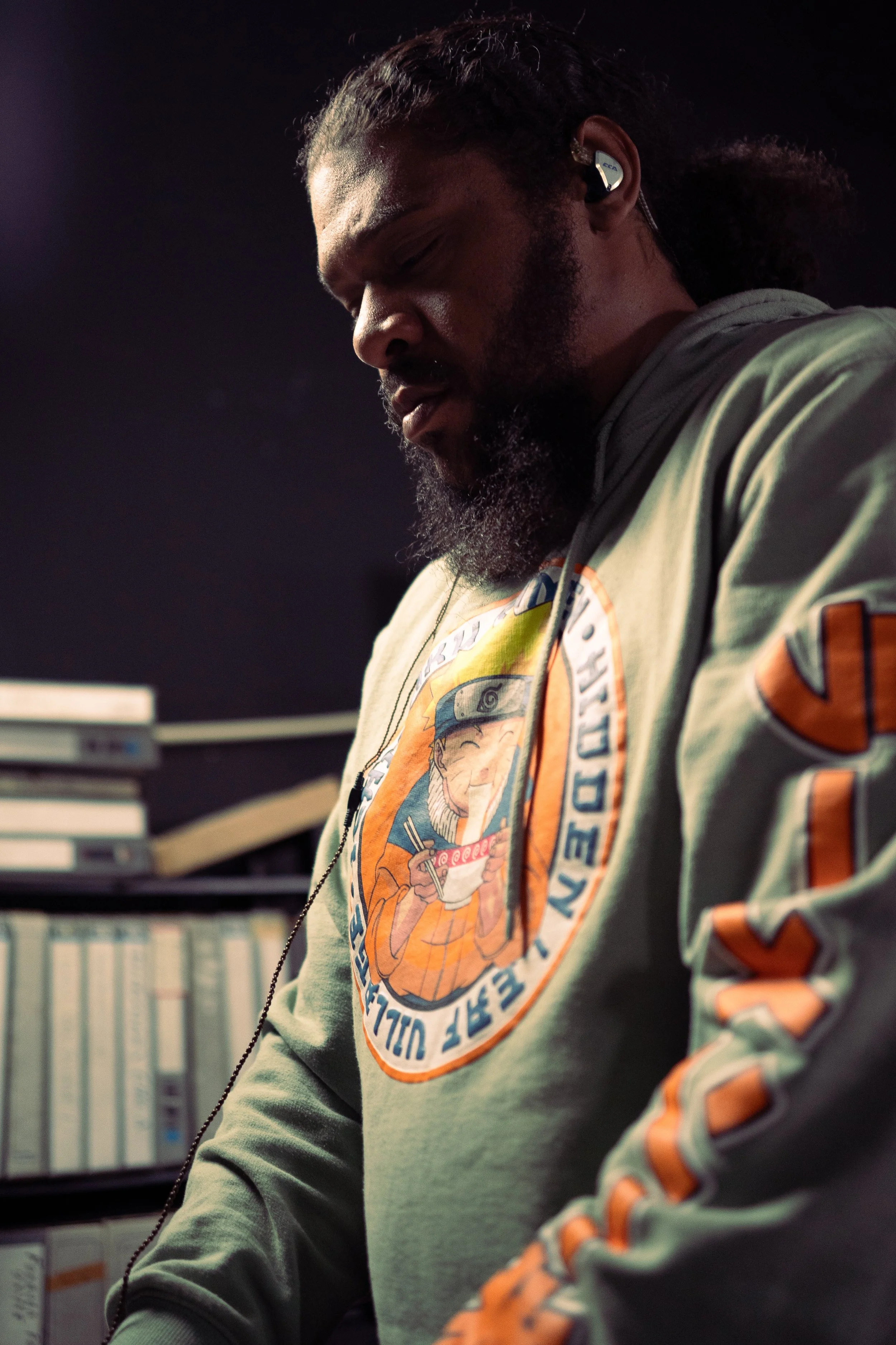 A man with a beard and long hair, wearing headphones and a gray sweatshirt with Naruto and Japanese characters on it, looking down in a library.