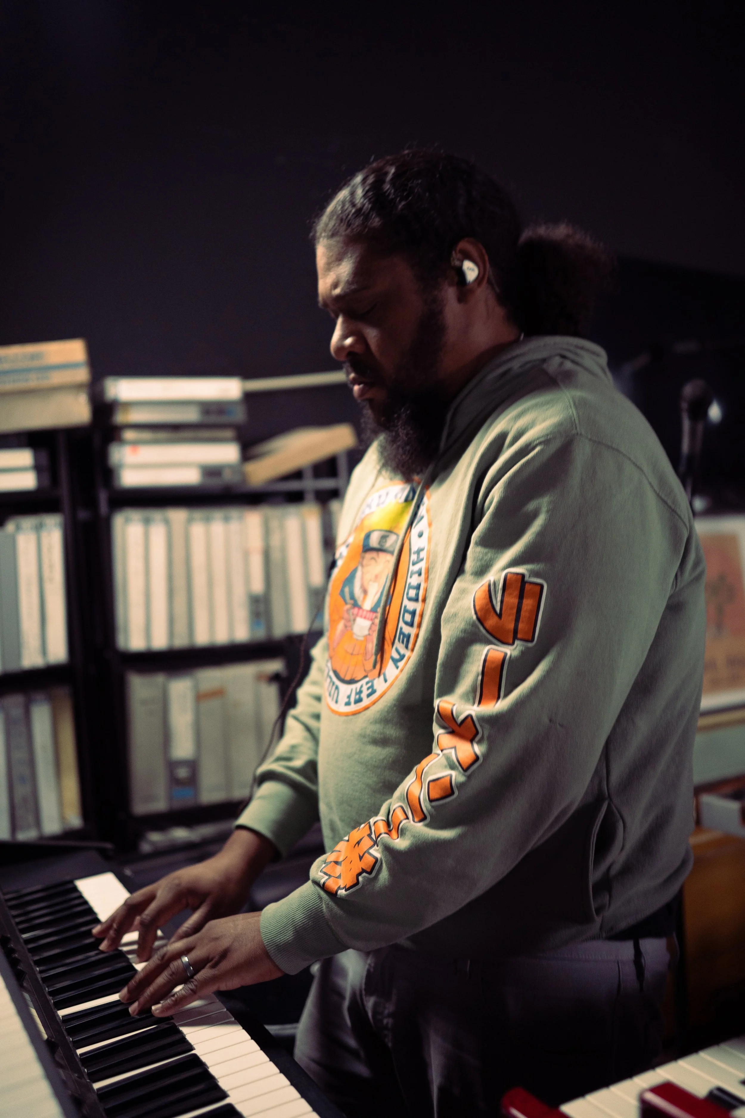 A man playing a keyboard in a dimly lit room, wearing a hoodie with a graphic logo on the front and patch on the sleeve, and wireless earbuds.