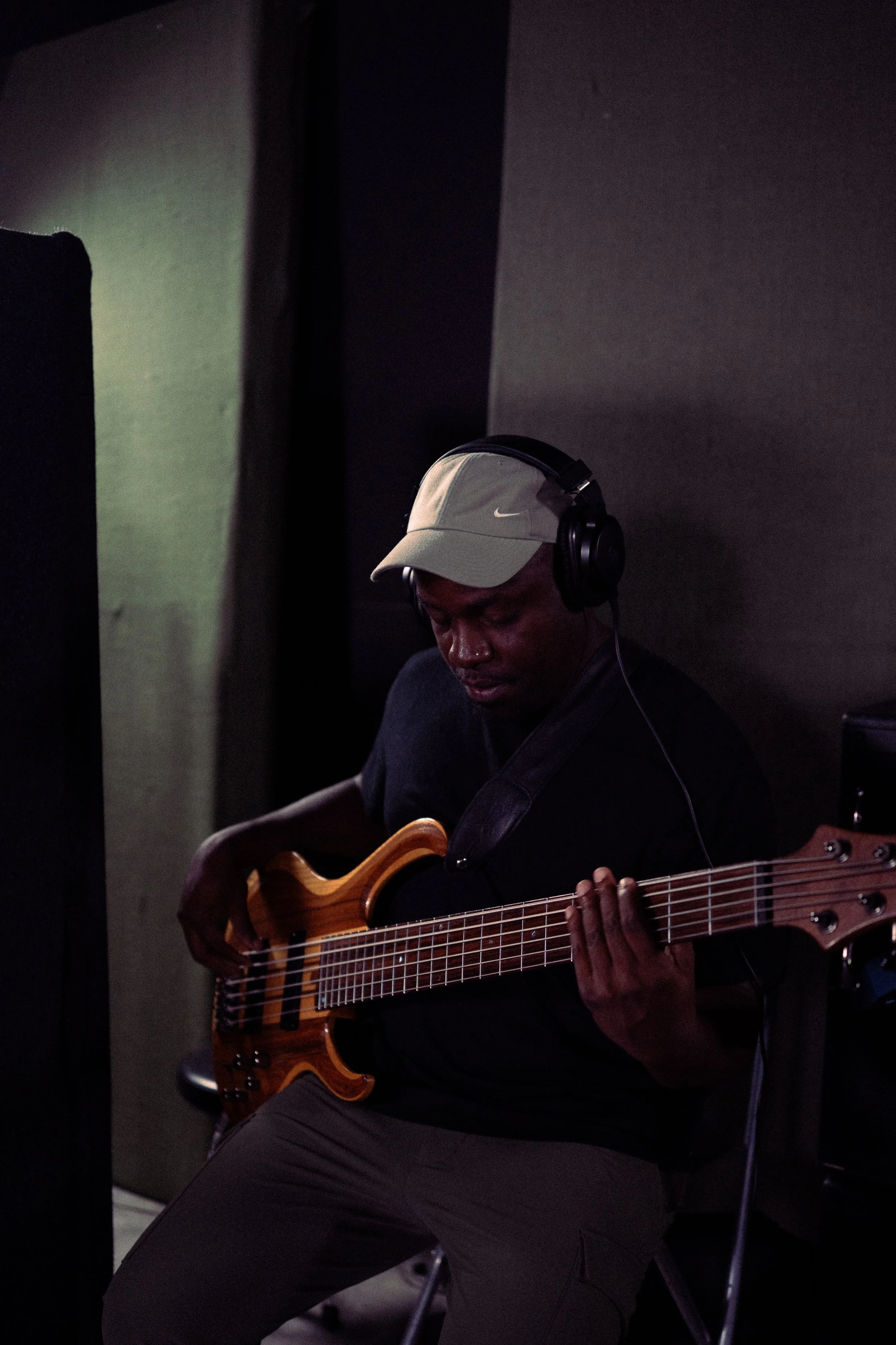 Man wearing a light-colored Nike cap and headphones playing a bass guitar in a dimly lit room.