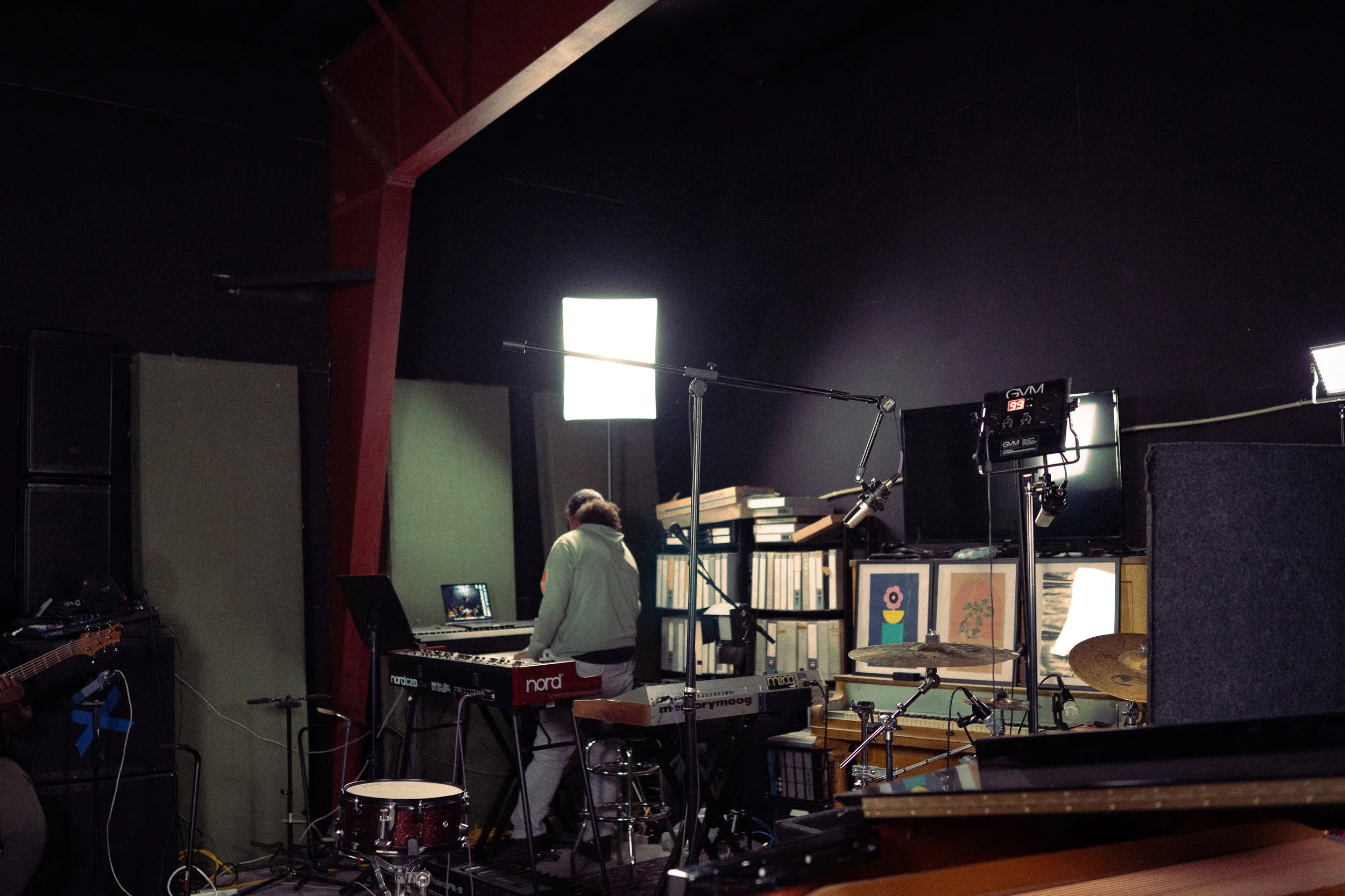 Music studio with keyboard, drums, microphone, and music equipment, with a person working at the keyboard.