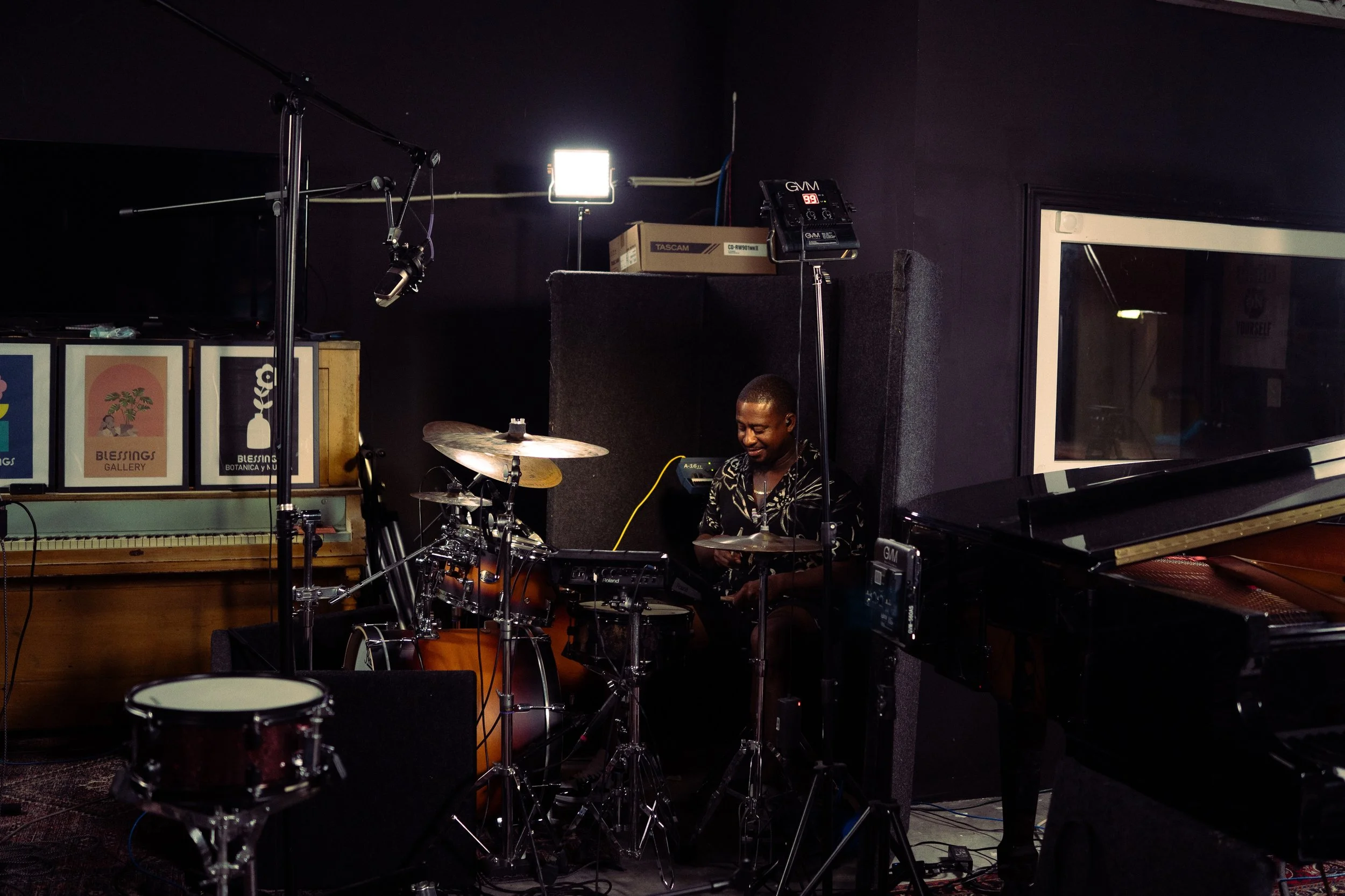 A man sitting at a drum set in a music studio, surrounded by musical equipment and a piano.