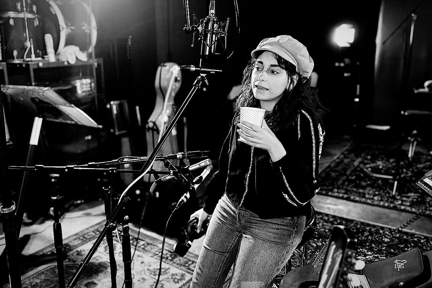 A woman with curly hair, wearing a beret and a black jacket, is holding a cup with a zigzag pattern and a camera while in a recording studio surrounded by musical instruments.