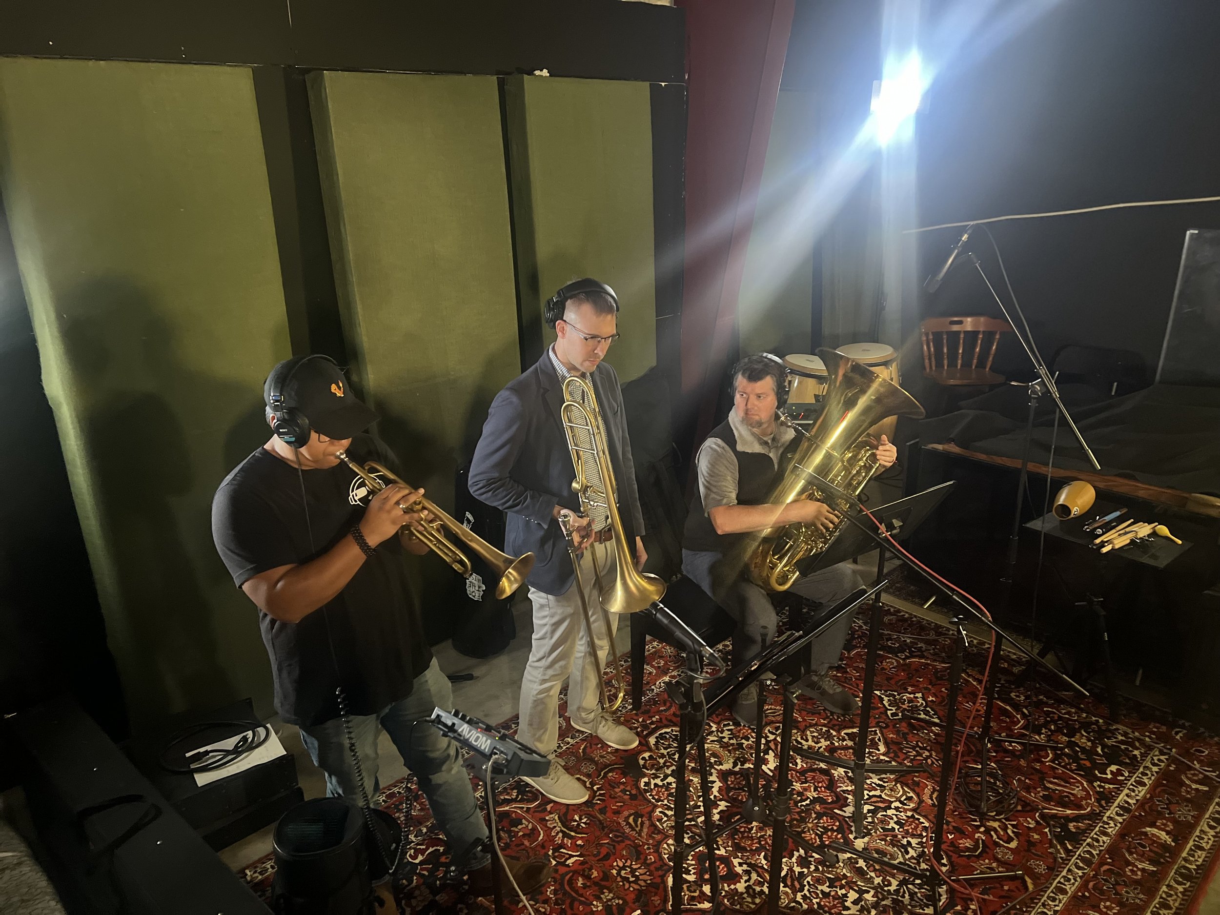 Three musicians in a recording studio playing brass instruments, including a trumpet, trombone, and tuba, with music stands and recording equipment around them.