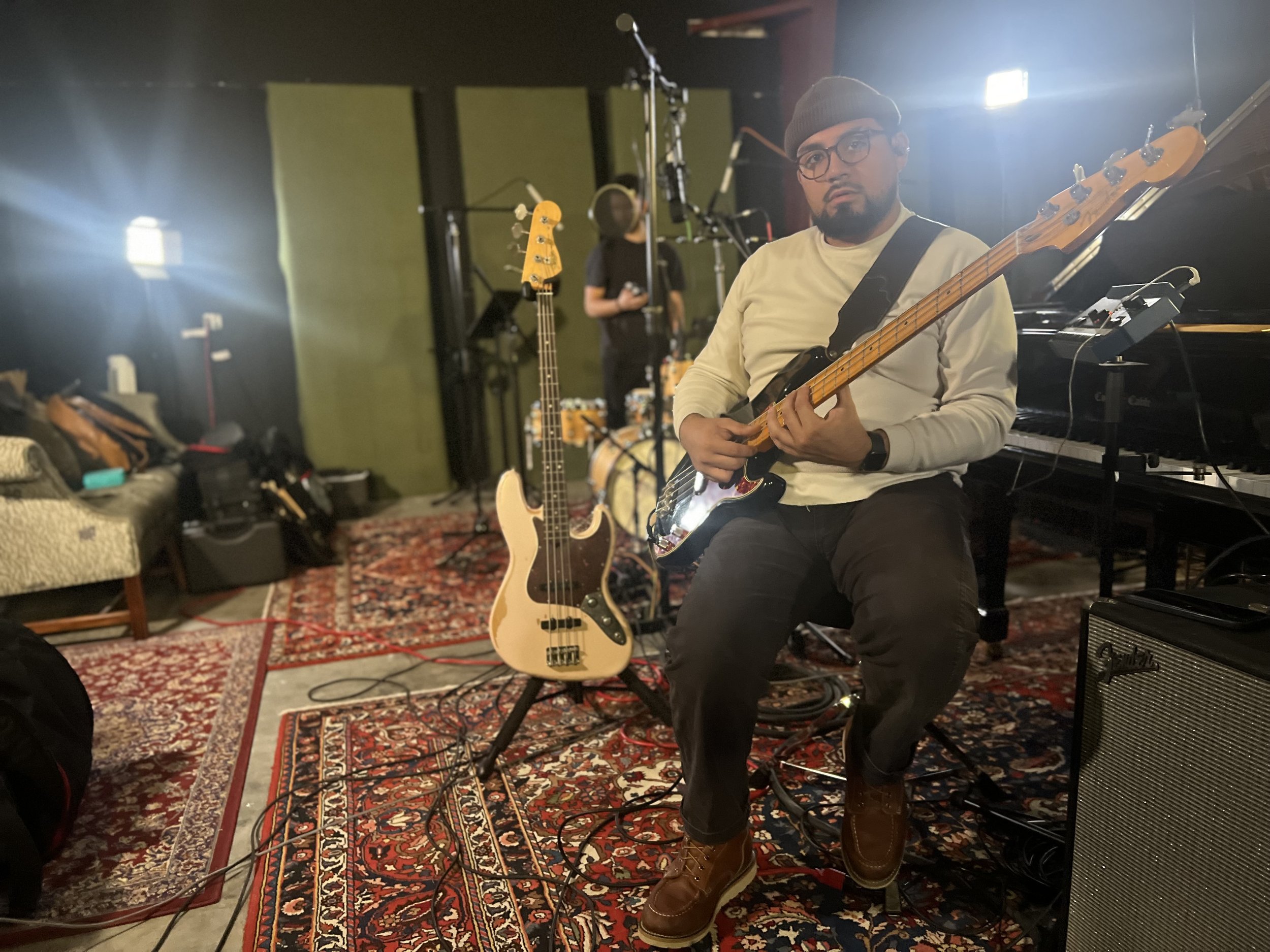 Musician sitting on a stool in a recording studio, playing an electric guitar. An electric bass guitar is standing on a stand next to him, and a drum set and keyboard are visible in the background. The studio has a patterned rug and walls with green