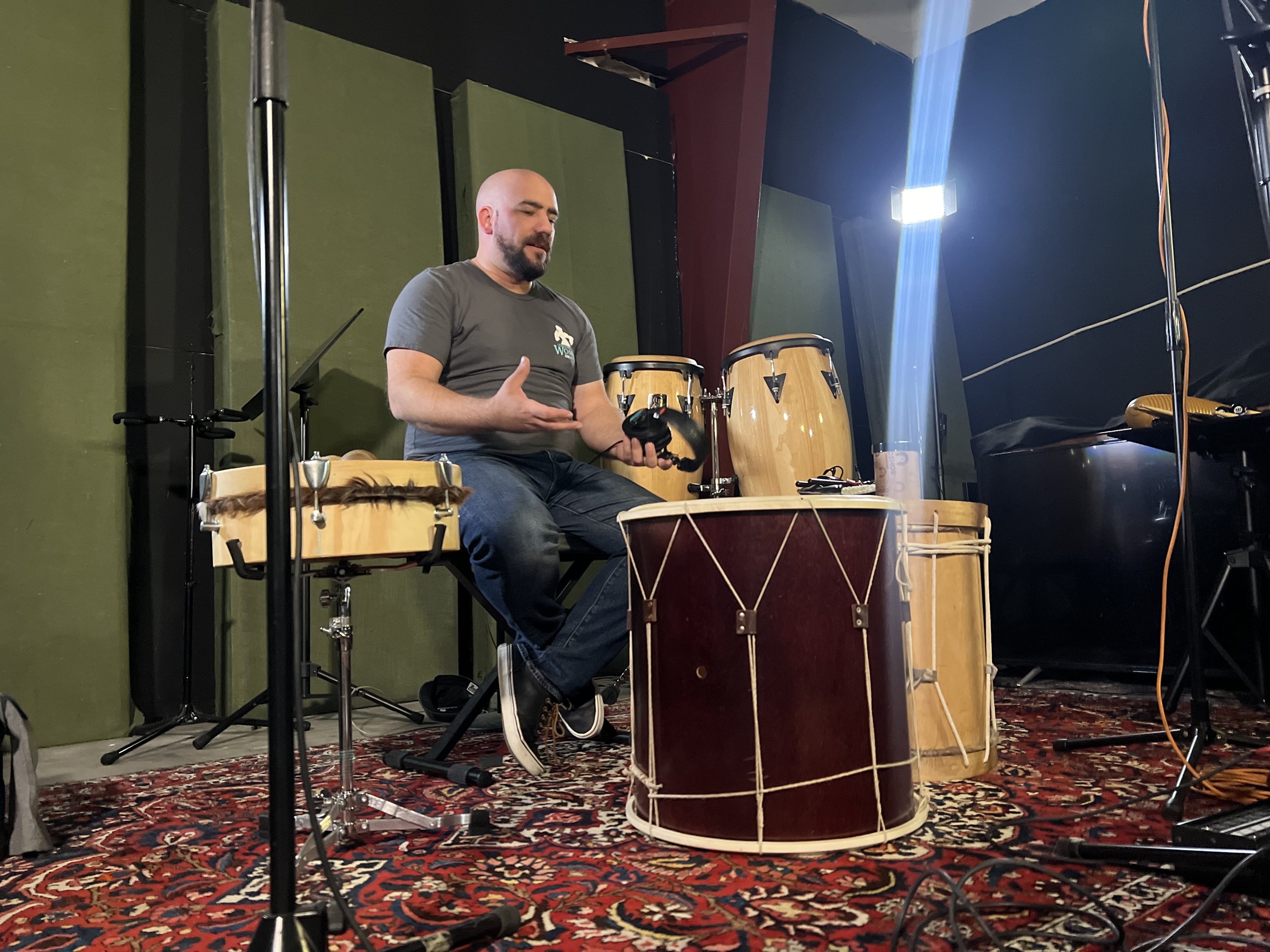 A man sitting on a chair holding a drumstick and a percussion instrument in a music rehearsal studio with various percussion instruments around him, including drums, congas, and a piano.