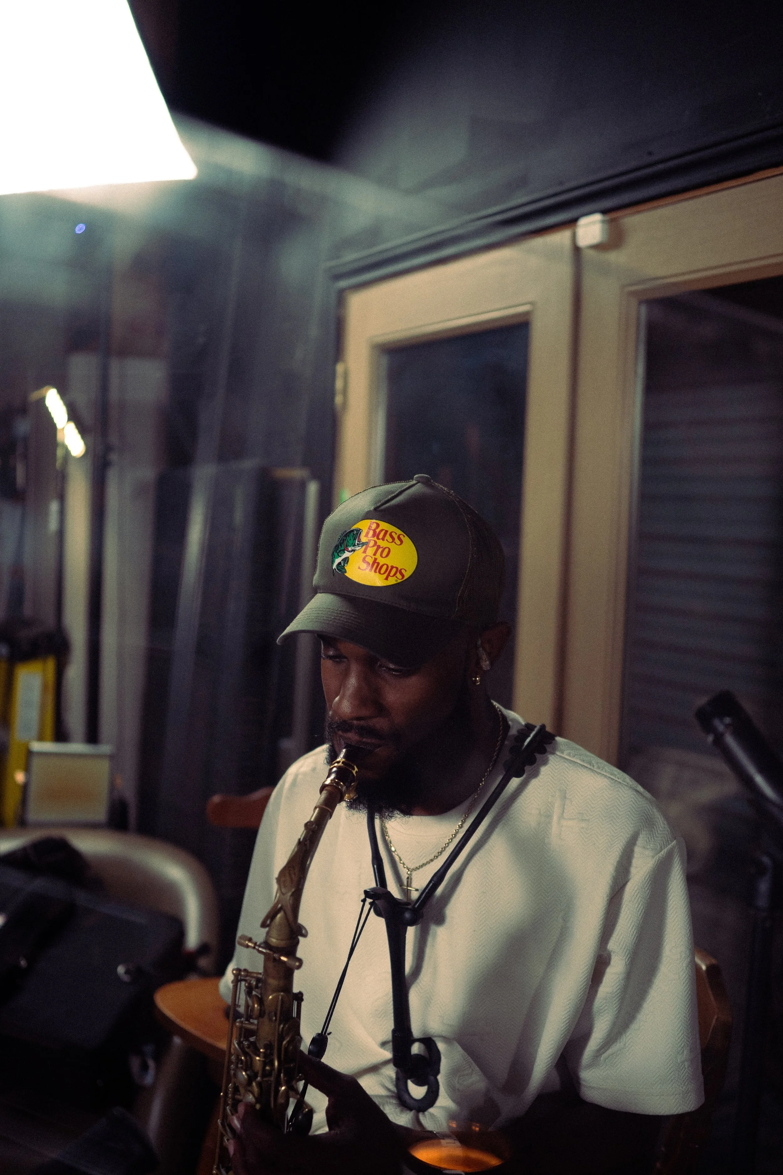 A man playing a saxophone indoors, wearing a Bass Pro Shops cap and white shirt, with music equipment around him.