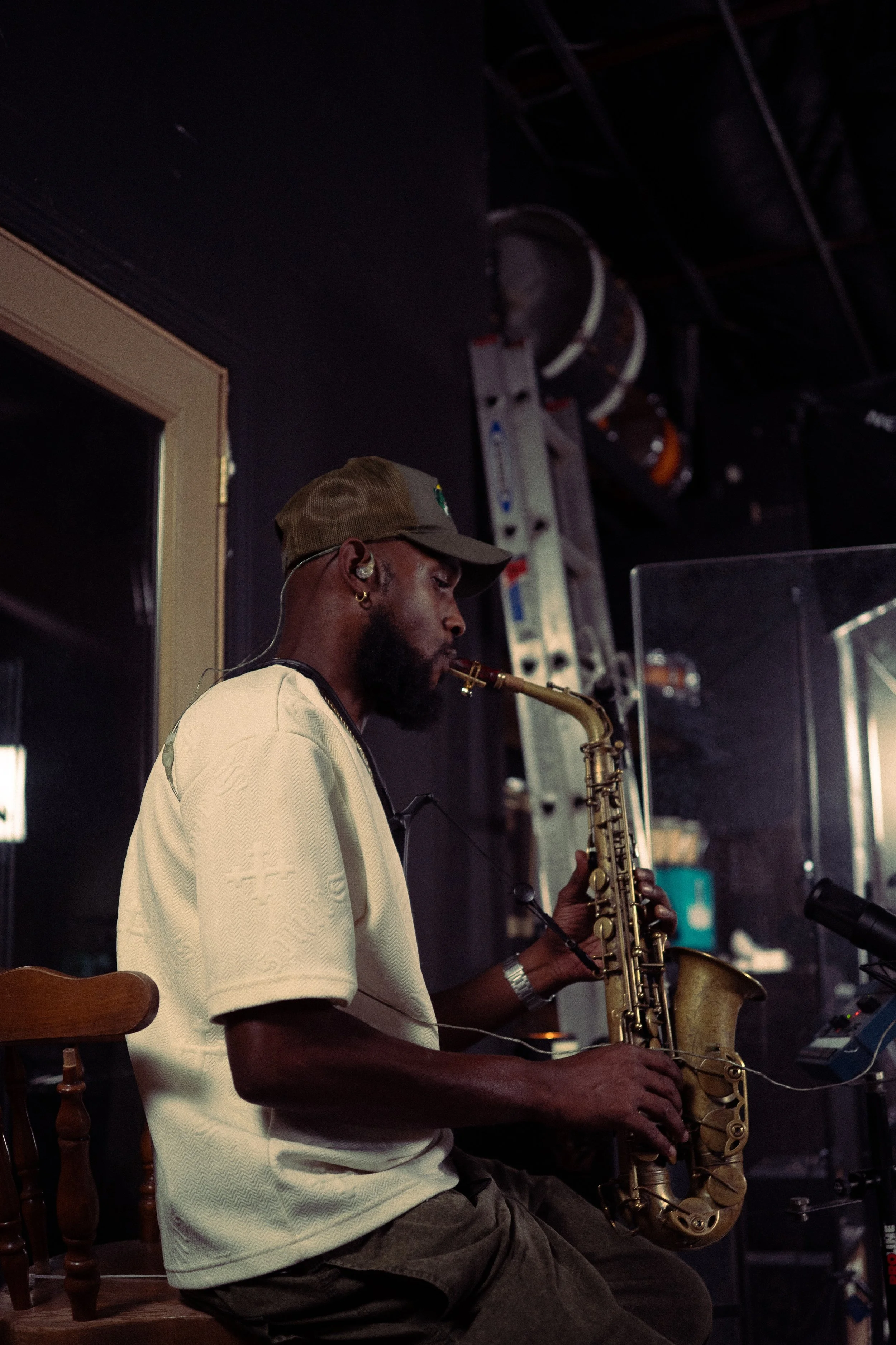 A man playing a saxophone on stage, wearing a beige shirt, a brown cap, and in-ear monitors.