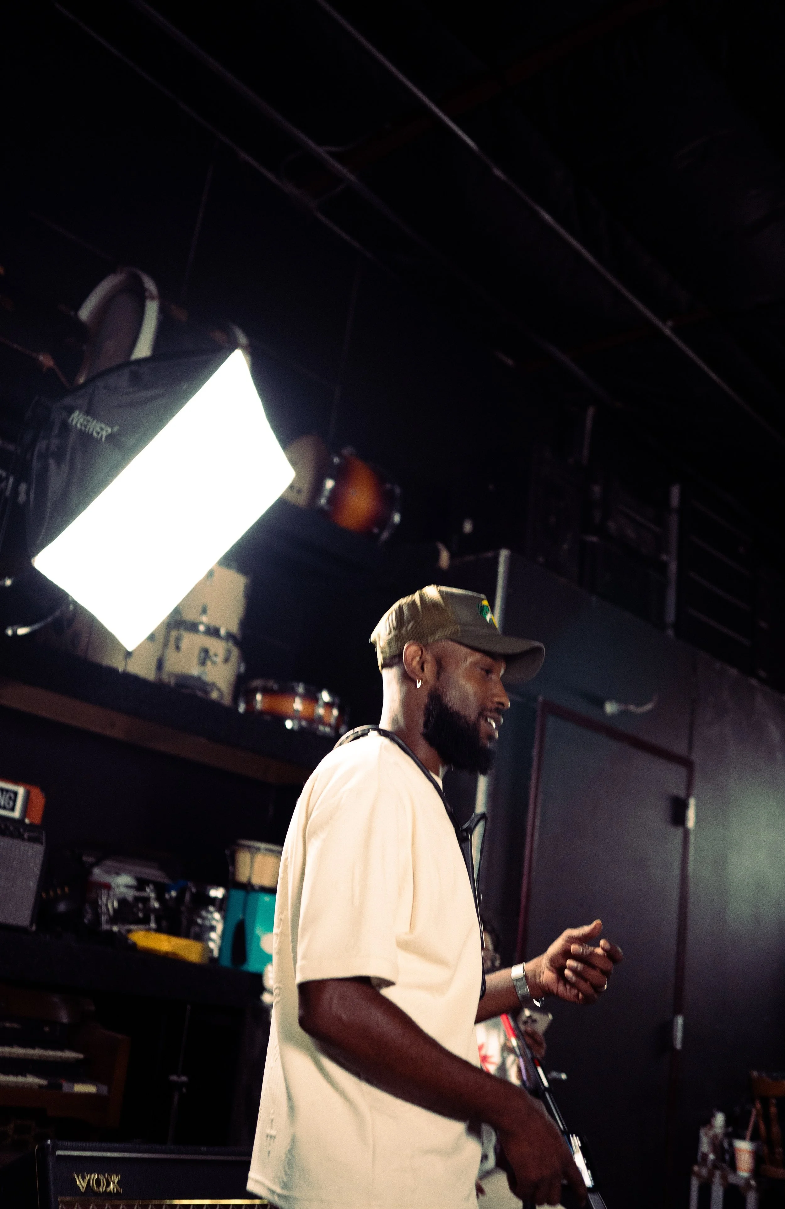A man with a beard wearing a beige shirt, cap, and earring, standing in a music studio or rehearsal space with musical instruments and equipment in the background and a large bright light to his left.