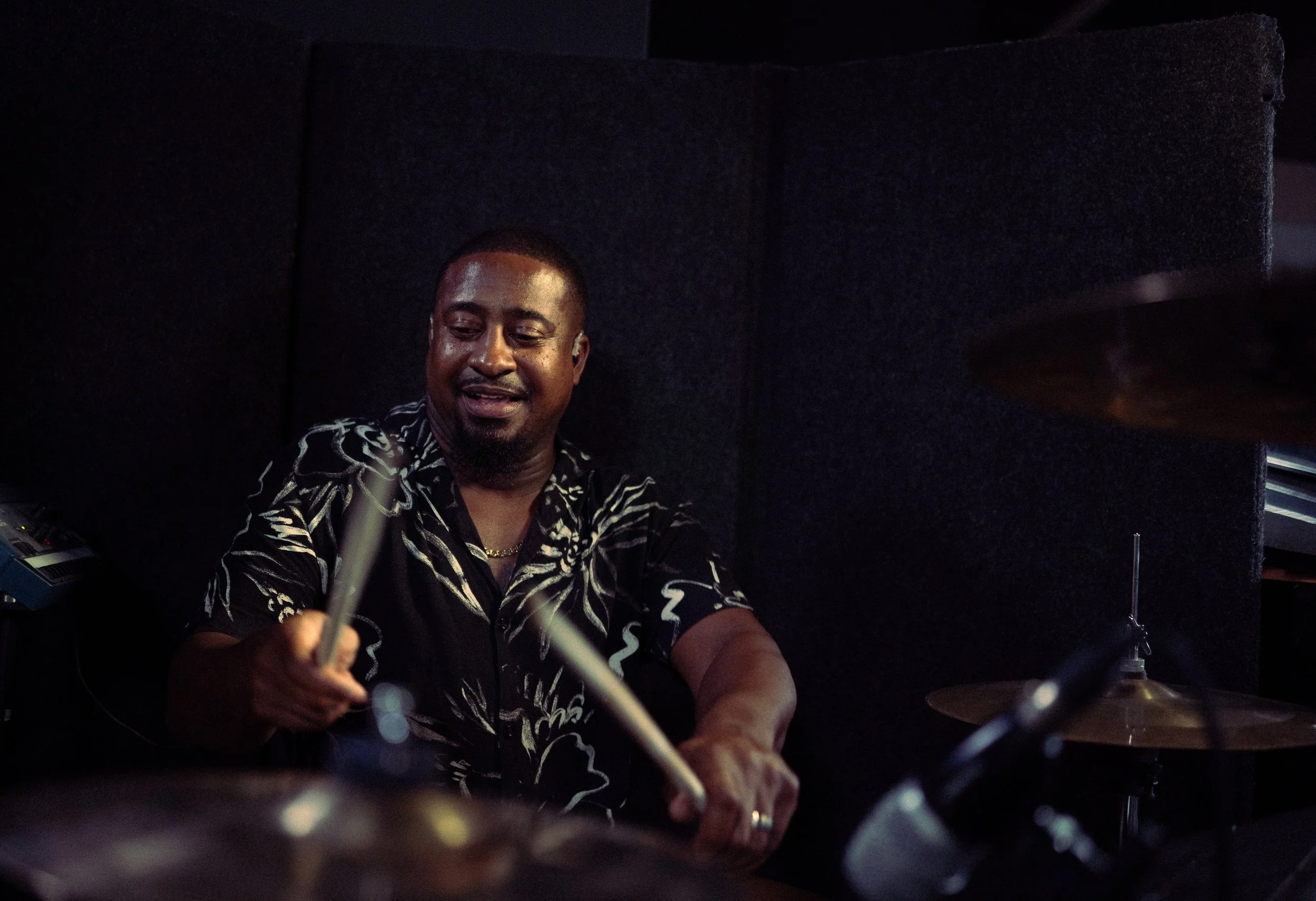 A man playing drums in a dark room, wearing a black shirt with white patterns, smiling, with drumsticks in his hands.