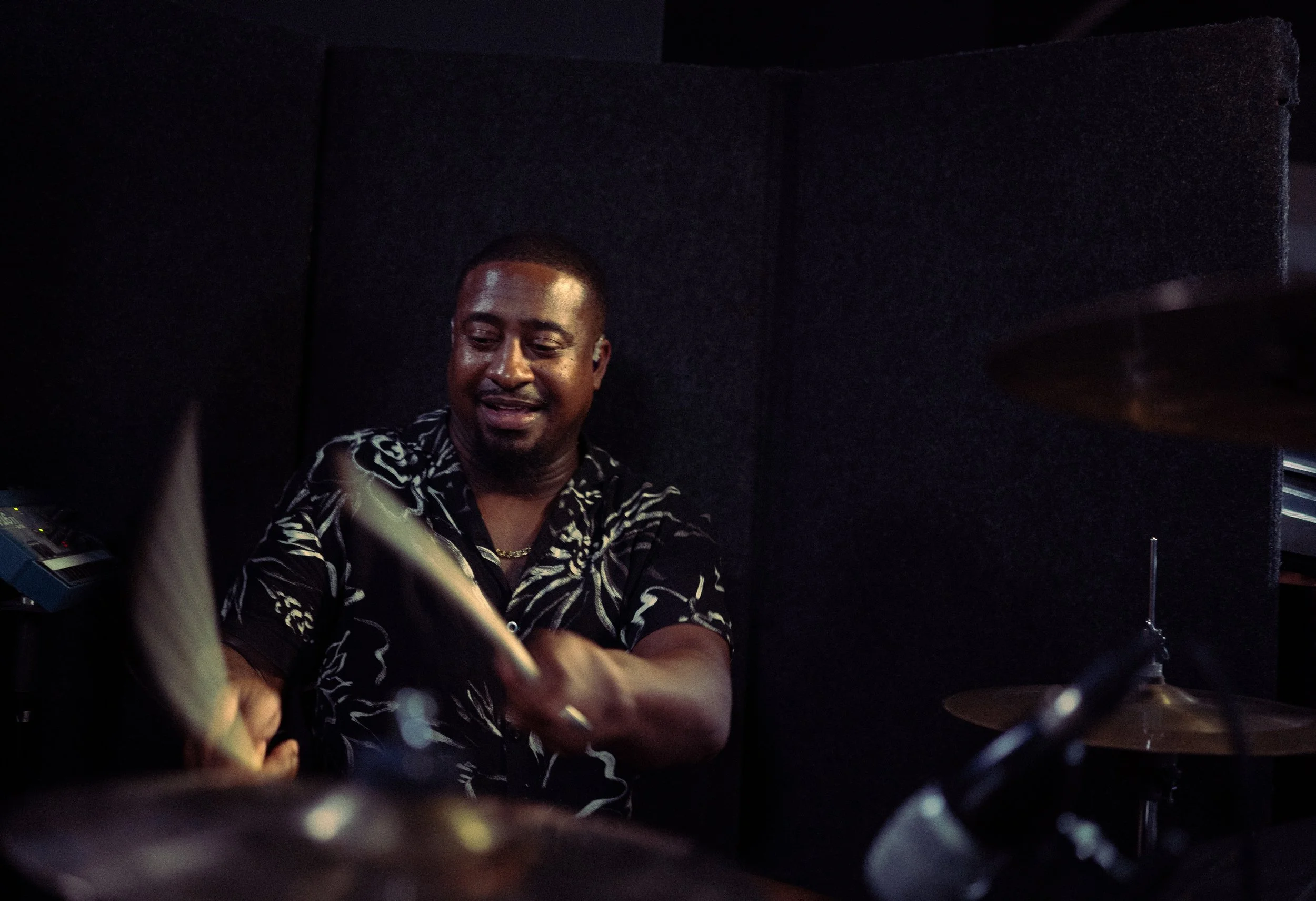 A man playing drums in a dark room, smiling and wearing a patterned shirt.