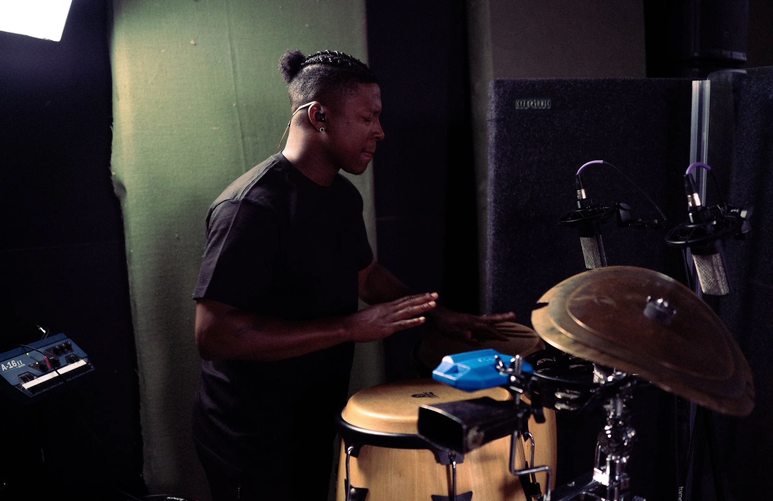 A person with dark hair styled in a bun, wearing all black, playing percussion instruments in a music studio.
