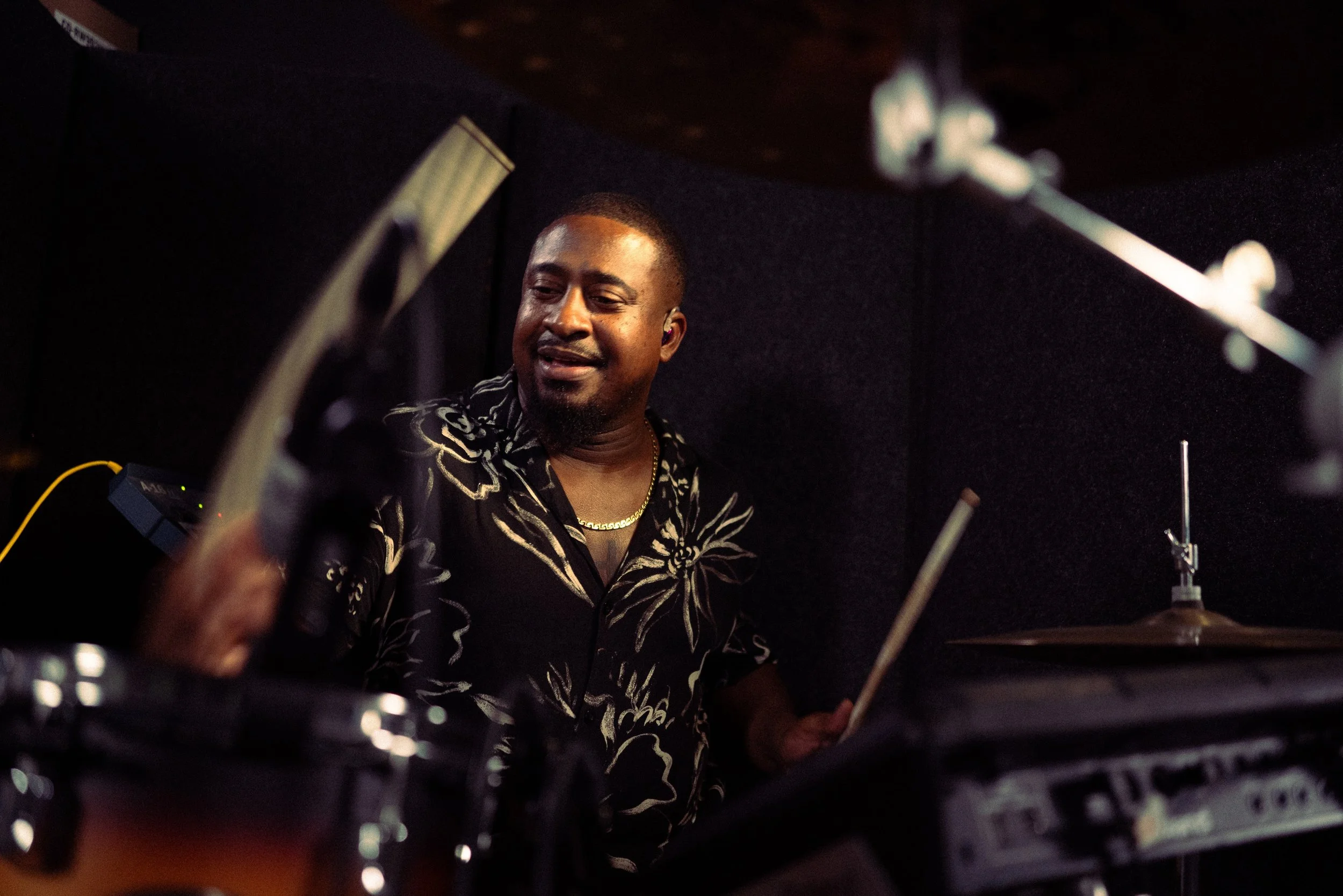 A man playing a drum set in a recording studio, wearing a black shirt with white floral patterns and a gold chain, smiling while looking at his instruments.