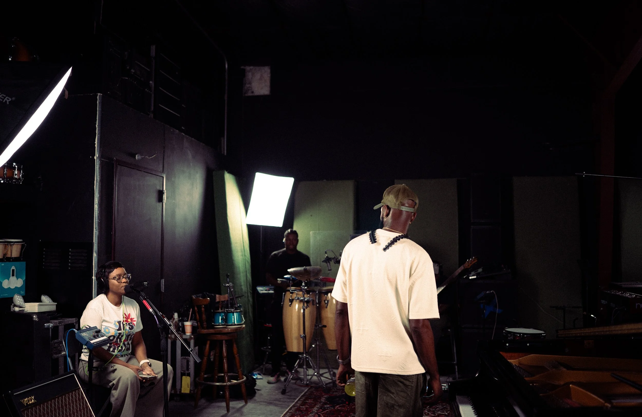 Musicians preparing in a dimly lit music studio with instruments, a woman with headphones sitting at a microphone, and a man with a cap facing away.