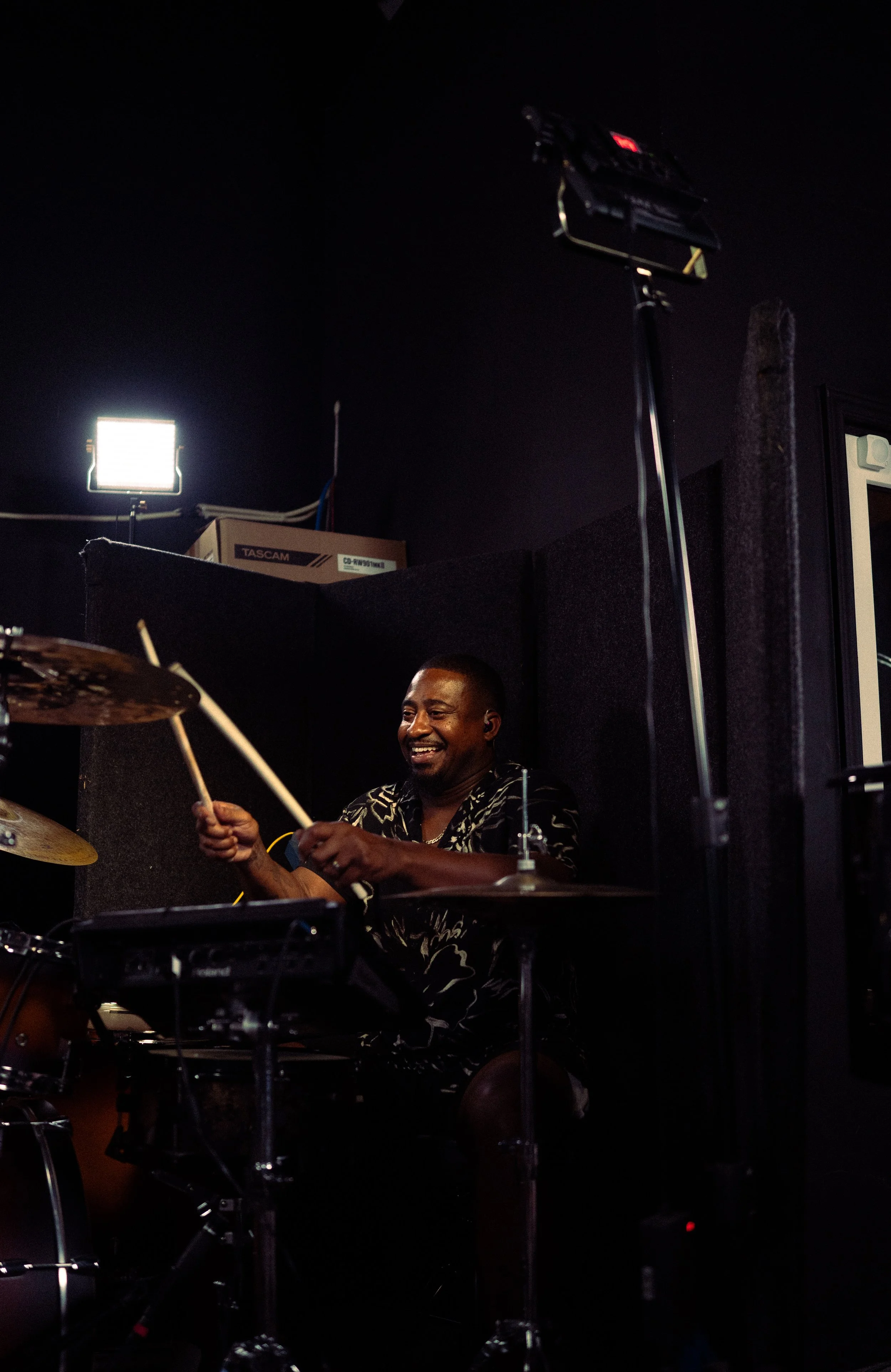 A man smiling while playing drums in a dark room with lighting equipment overhead.