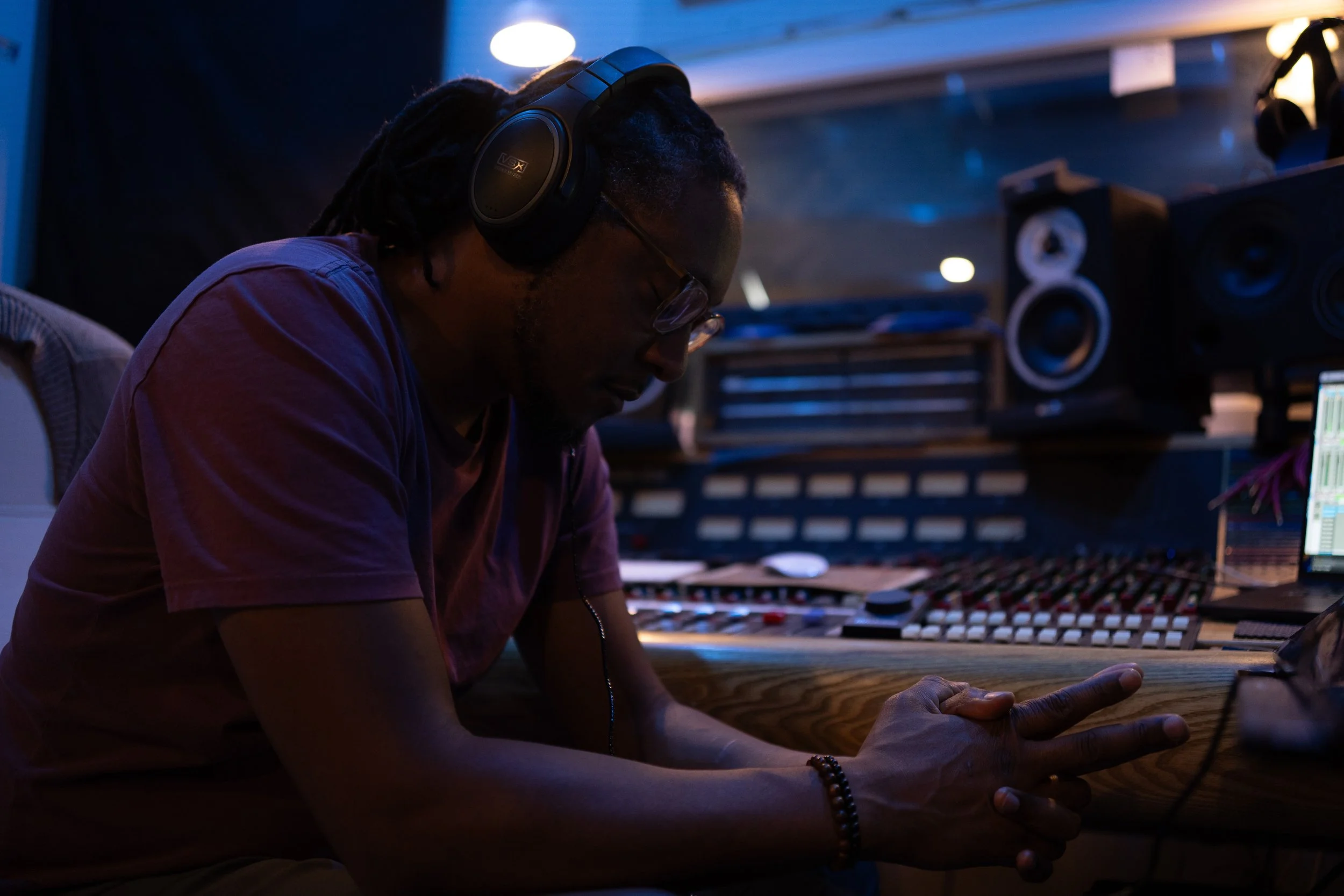 A man wearing headphones and glasses sitting in a recording studio, with music equipment and a computer screen visible on the desk.