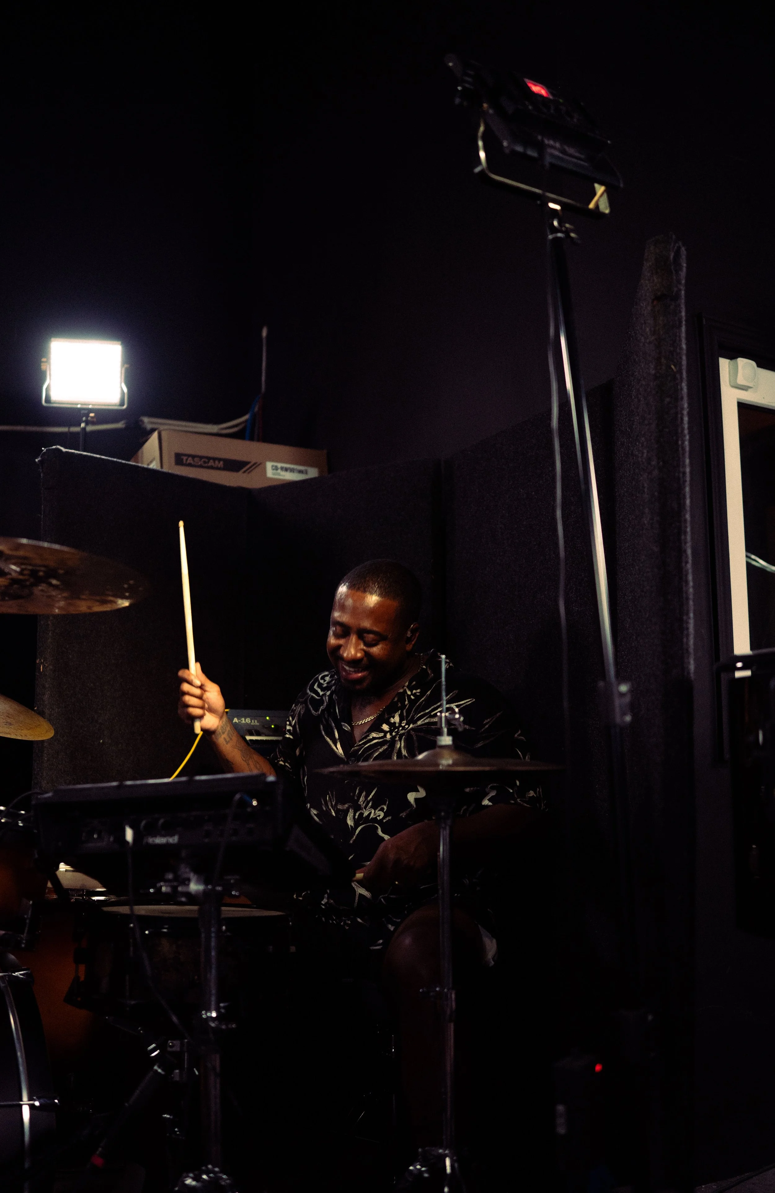 A man smiling and playing drums in a dimly lit room with stage lighting equipment.