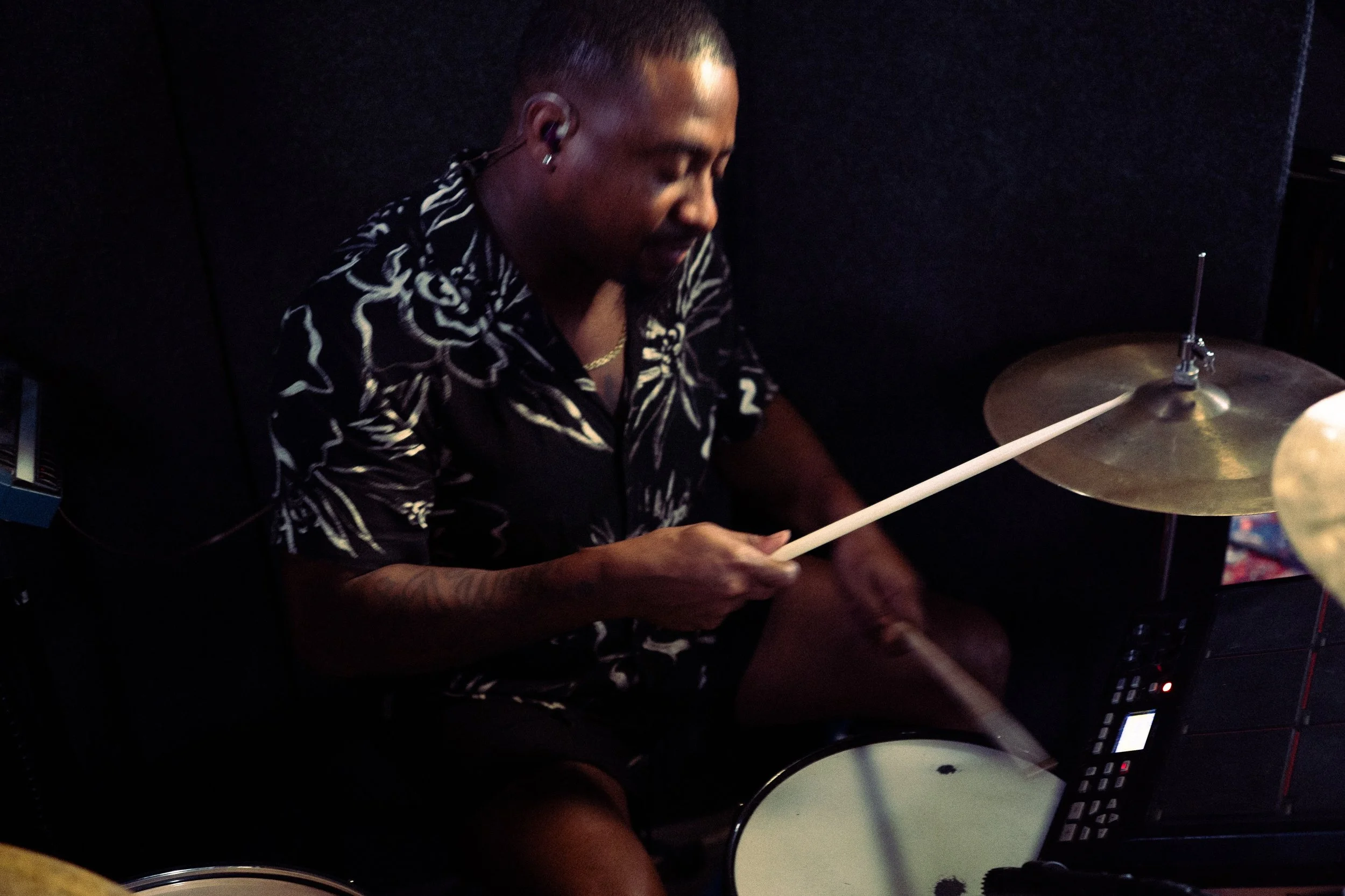 Person playing electronic drum set, wearing a black and white patterned shirt, in a dimly lit room.