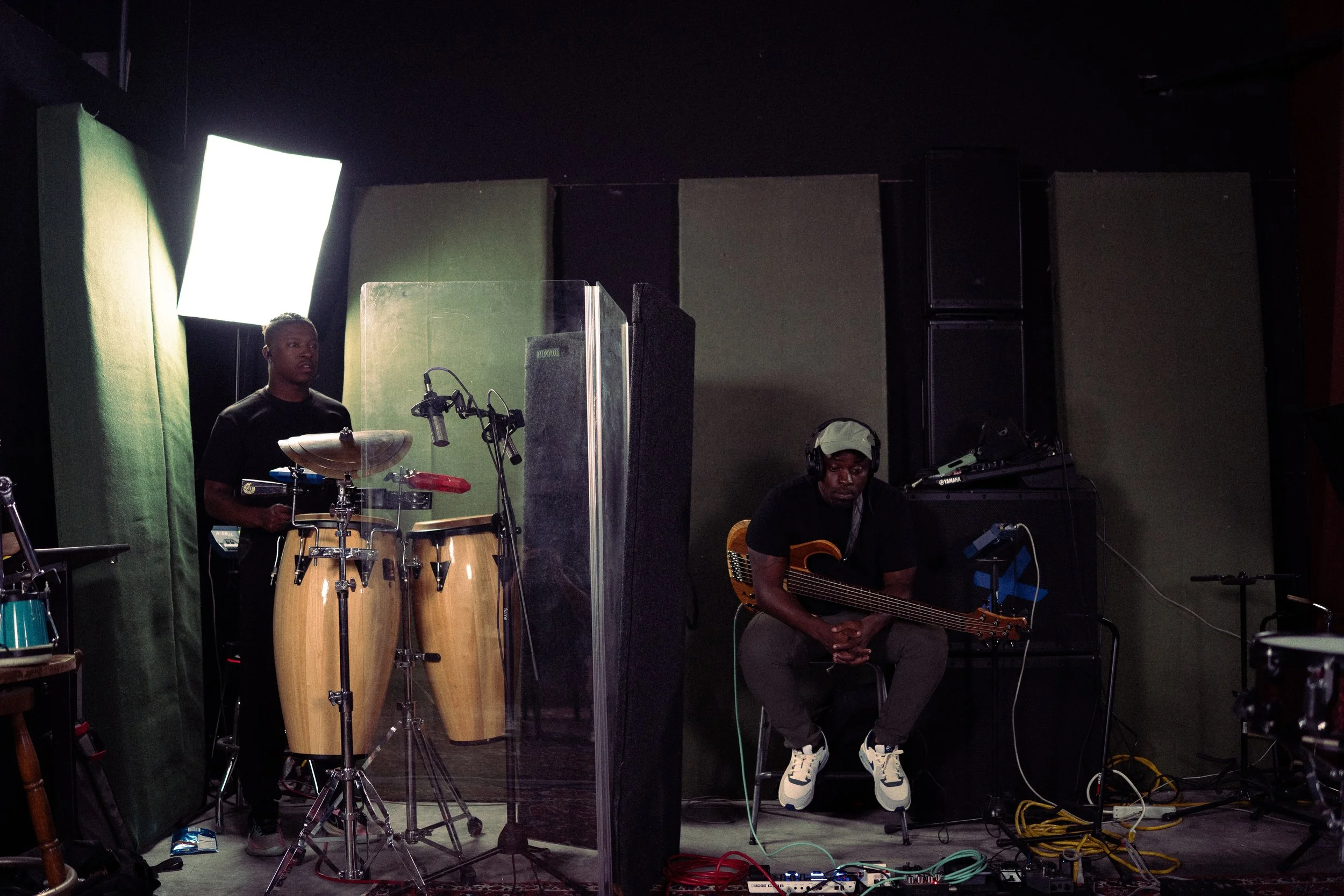 Two musicians in a recording studio: one standing behind percussion instruments, the other seated with a guitar, with professional audio equipment around.